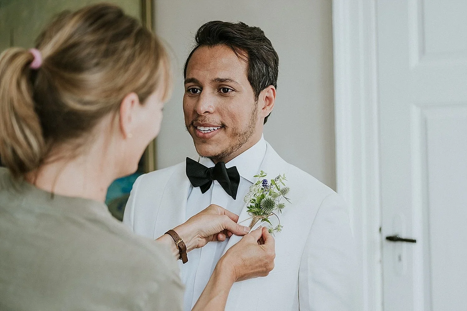 A gay groom in a white tuxedo and black bowtie having a summer boutonniere with white and blue flowers pinned to his lapel by wedding planner and founder Louise before the ceremony at The Old Merchant's House.