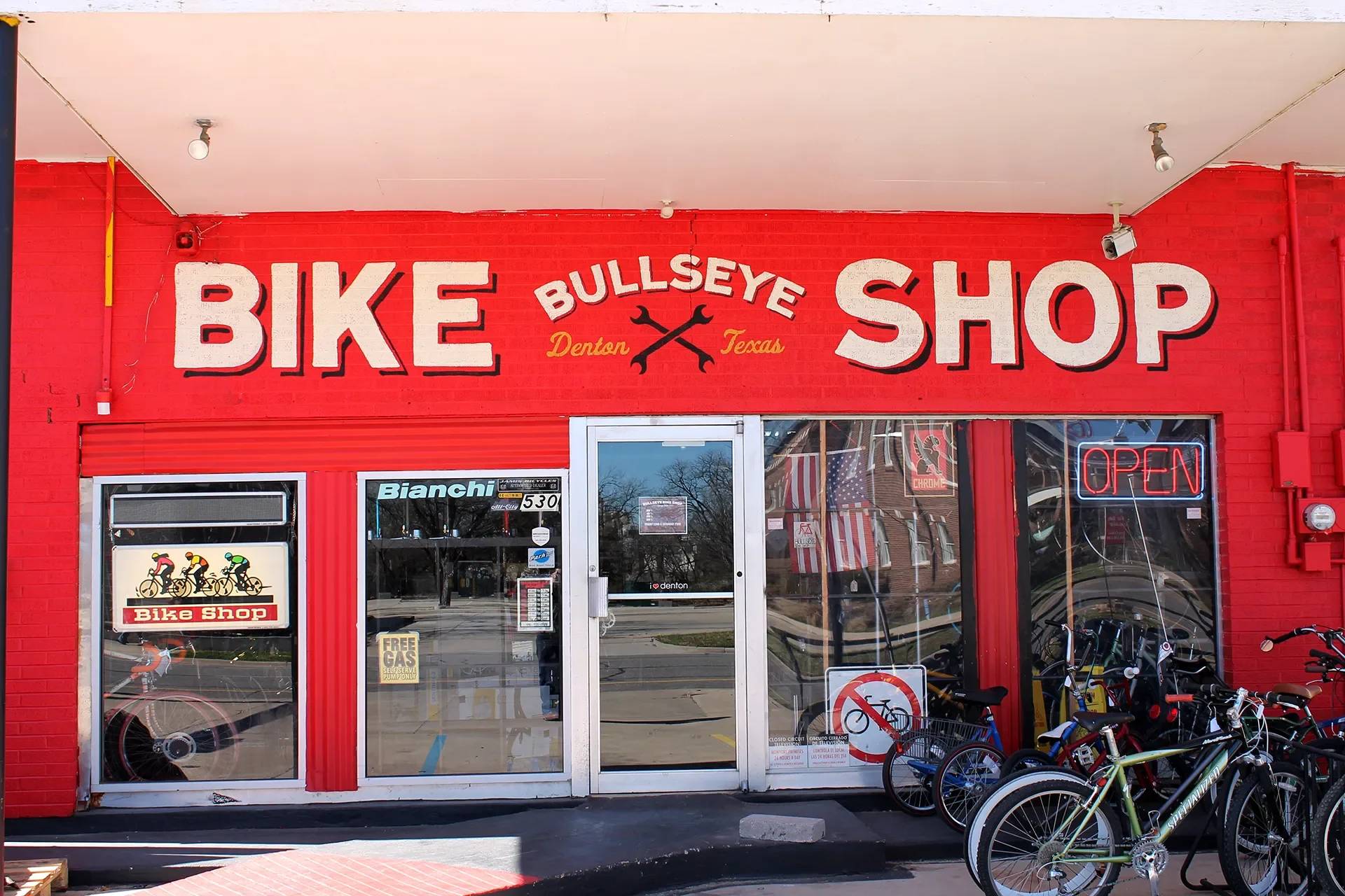Red brick exterior of a bike shop named 'Bullseye Bike Shop' in Denton, Texas with bicycles outside and an illuminated 'Open' sign in the window.