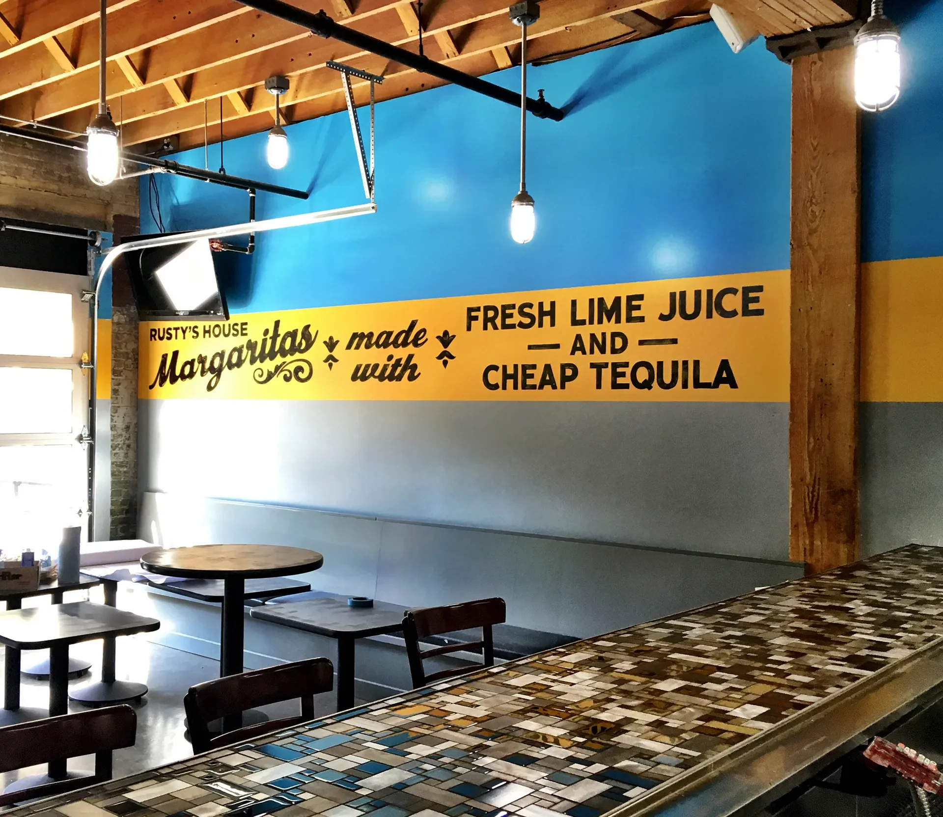 Interior of a casual restaurant with a large yellow and gray wall sign that reads 'Rusty's House Margaritas made with fresh lime juice and cheap tequila.' There are wooden tables and chairs, a mosaic-tiled counter, and exposed wooden beams on the cei