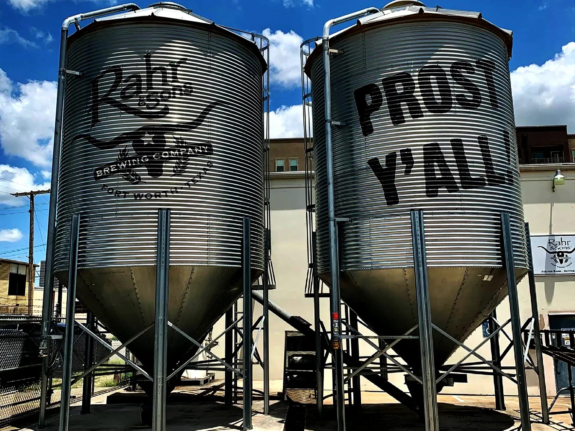 Two large cylindrical metal tanks with stenciled branding, one labeled "Rahr & Sons Brewing Company" and the other with the slogan "Prost Y'all". They are outdoors under a blue sky with scattered clouds.