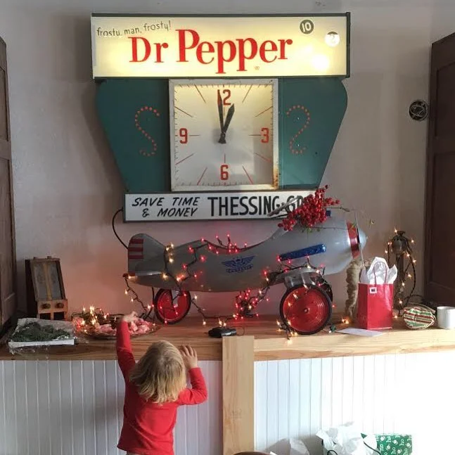 A vintage-style Dr. Pepper clock with red and turquoise colors hanging on a wall, decorated with Christmas lights and ornaments. Below the clock, a small child in a red shirt stands near a wooden counter, looking at festive decorations including a mo