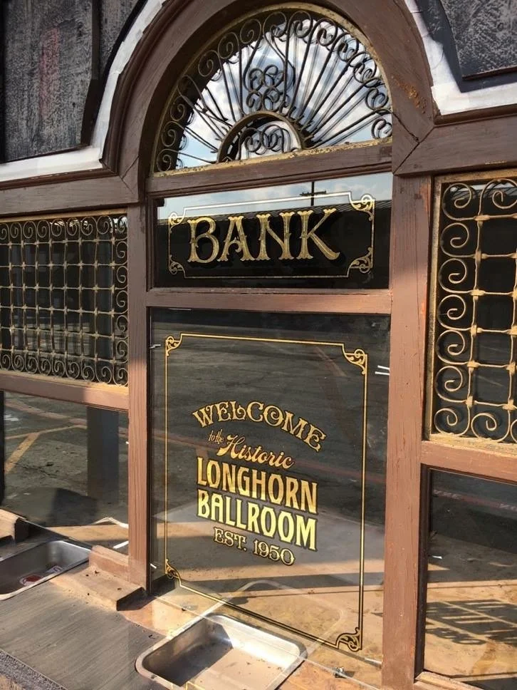A vintage-style street sign on a wooden structure with decorative ironwork, indicating the entrance to the Longhorn Ballroom, established in 1950, with a welcome message and the word 'BANK' at the top.