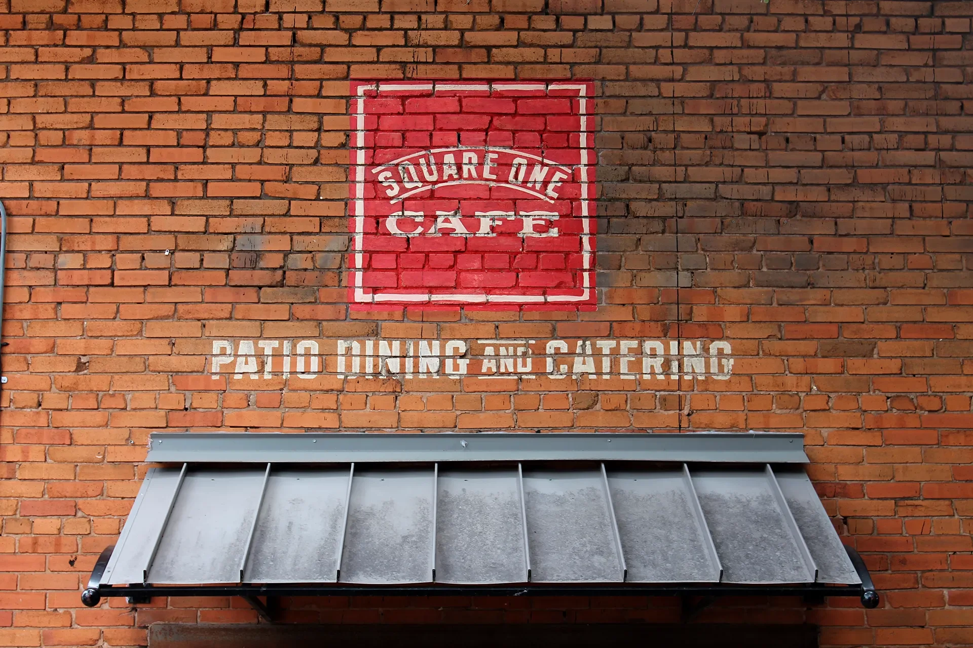 Brick wall with a red square sign reading 'Square One Cafe' and white text below stating 'Patio Dining and Catering', with a metal awning underneath.