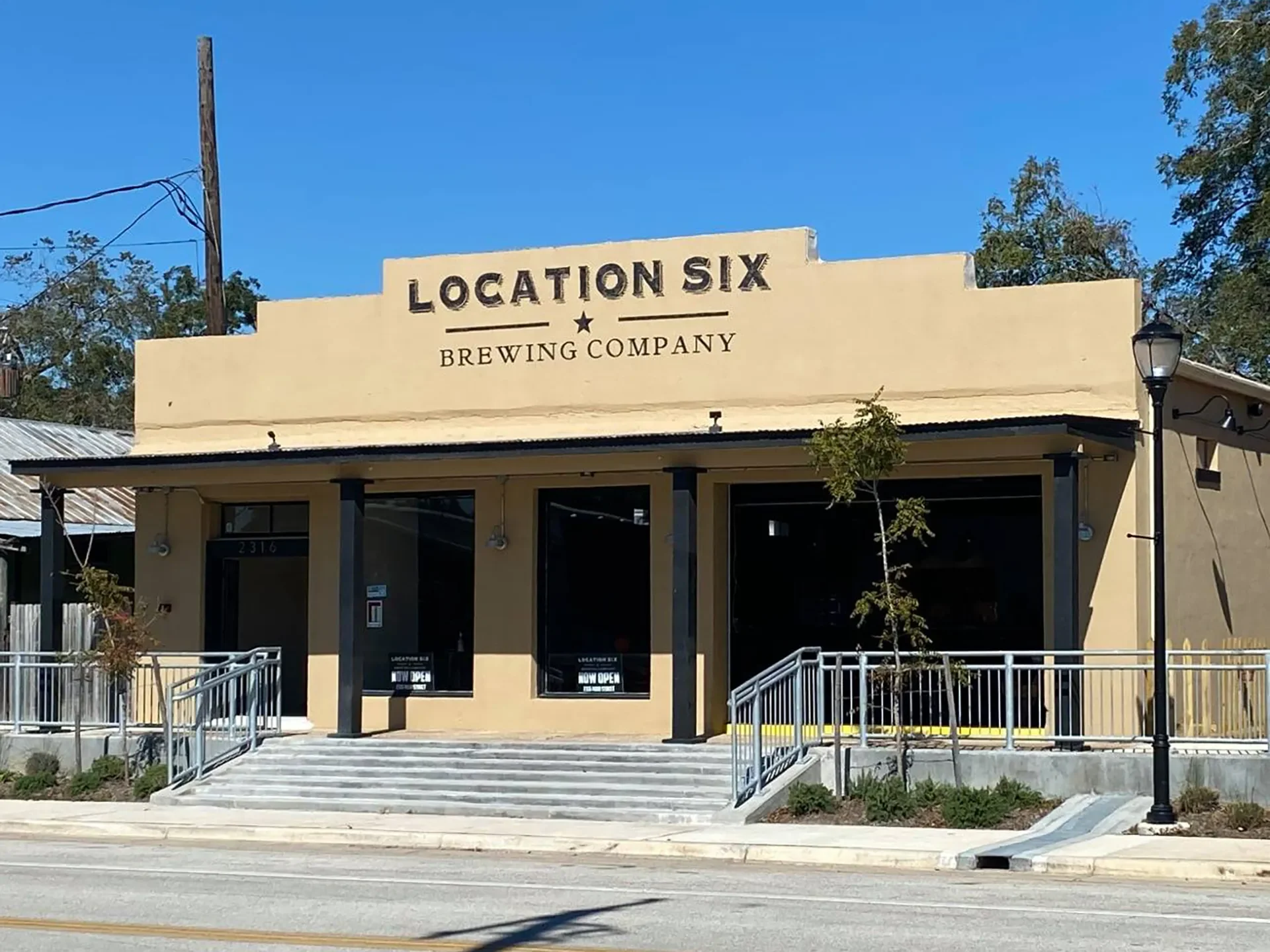 Front view of a beige building with a sign that reads 'Location Six Brewing Company' and 'Now Open'. It has stairs leading to the entrance, a ramp, and a street lamp in front.