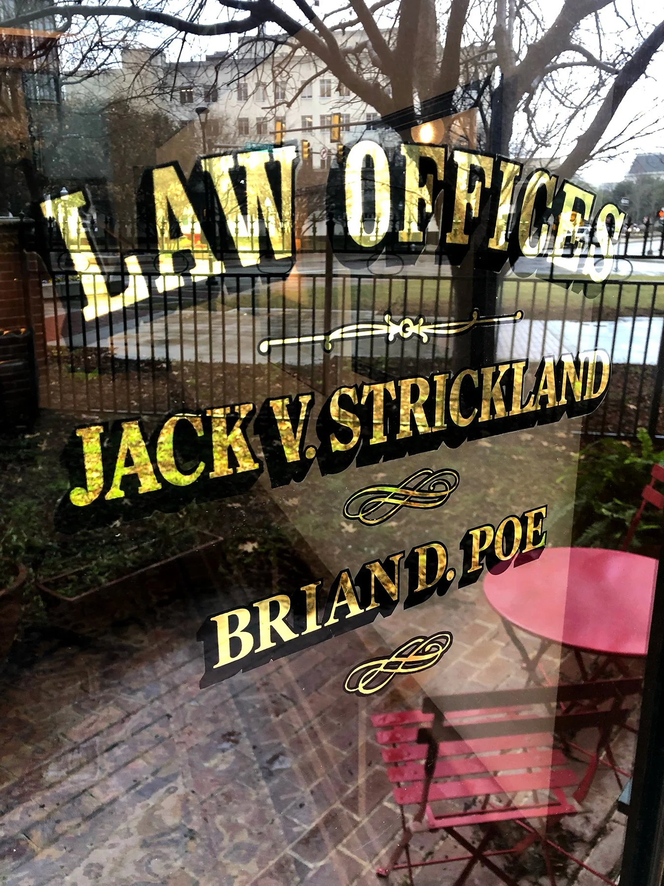 Reflected view of a patio with a red table and chairs seen through a window with the words 'Law Offices Jack v. Strickland' and 'Briand P.O.E.' written on it.