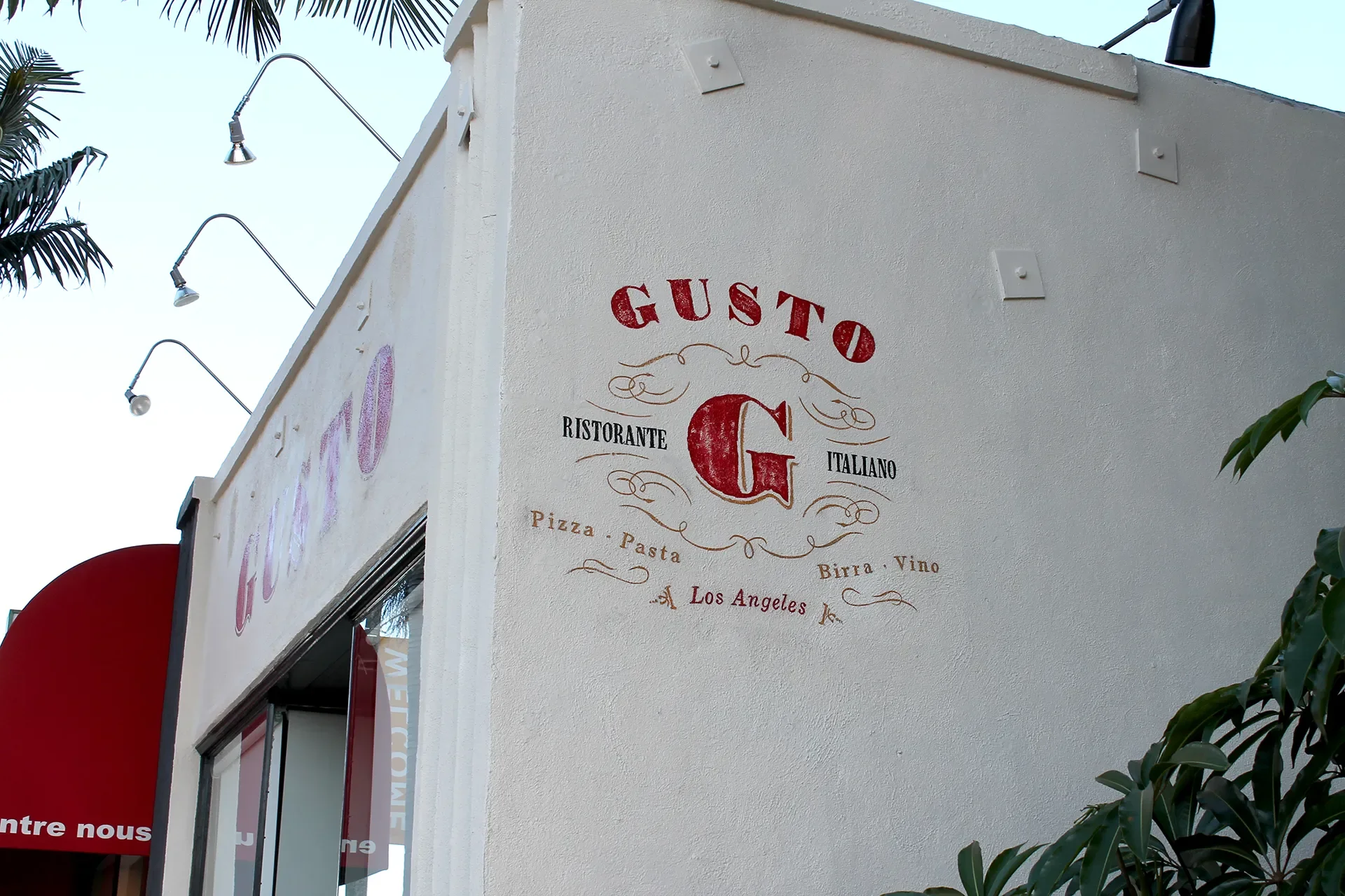 Exterior view of Gusto Italiano restaurant in Los Angeles with a white wall sign displaying the restaurant's name, type of cuisine, and offerings such as pizza, pasta, beer, and wine. There are several gooseneck lights mounted on the wall above, and 