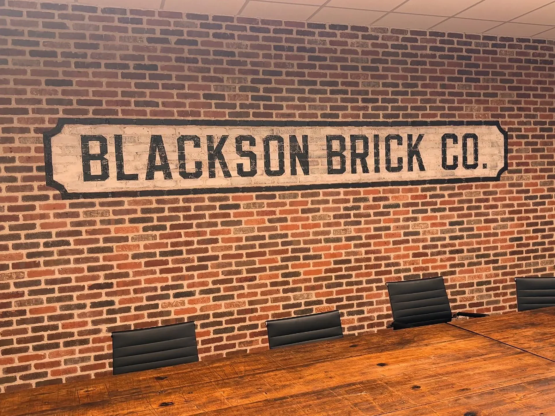 Interior of a conference room with a large brick wall featuring a vintage sign that reads 'BLACKSON BRICK CO.' in black text on a white background. There is a wooden table and black office chairs.