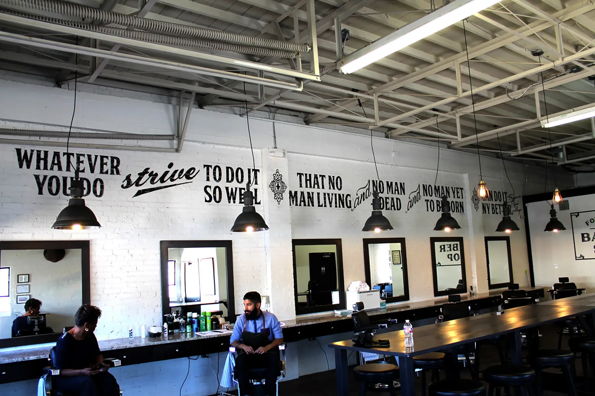 Interior of a modern barbershop with black pendant lights, white brick walls, and mirrors. A man with a beard and a woman are seated, and the wall features a quote painted in black text.