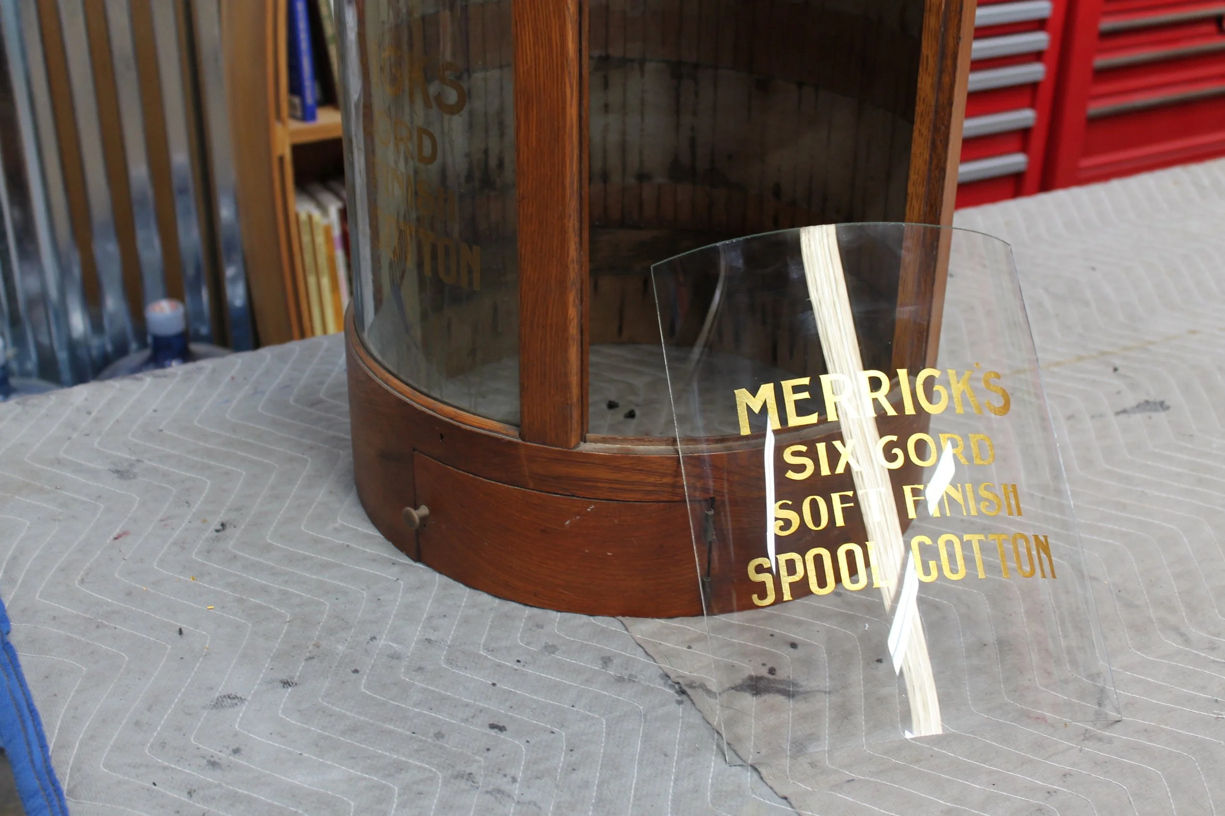 A woodworking project with a curved wooden frame and a clear plastic shield with gold text. The text reads 'MERRICK'S SIX GORD SOFT FINISH SPOOLED COTTON.' The project is on a cloth-covered work surface in a workshop.