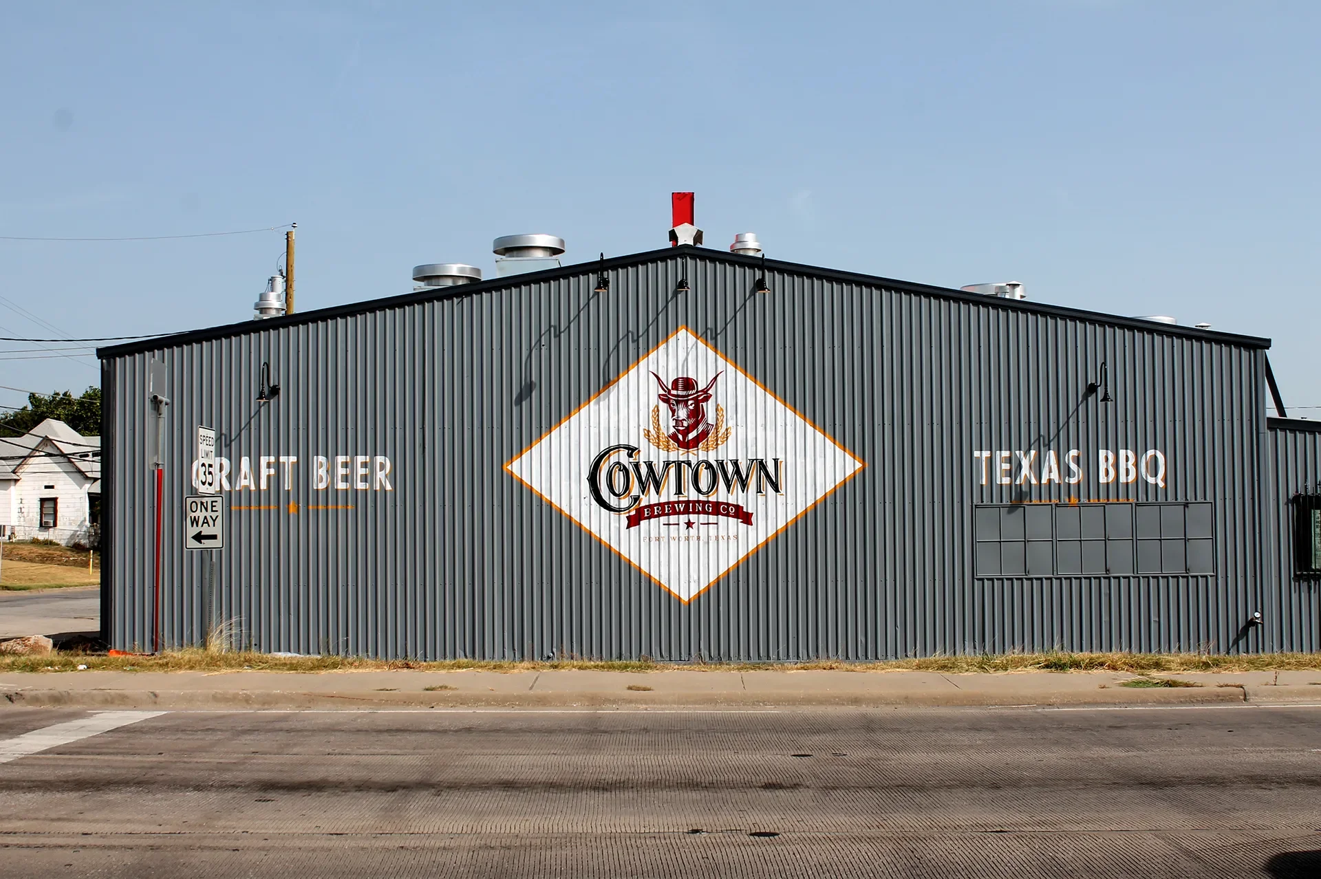 A large industrial-style building with gray corrugated metal siding. The wall features a logo of a cow wearing a cowboy hat and the name 'Cowtown Brewing Co.' in a diamond shape. To the left, the words 'Craft Beer' and a speed limit sign are visible.