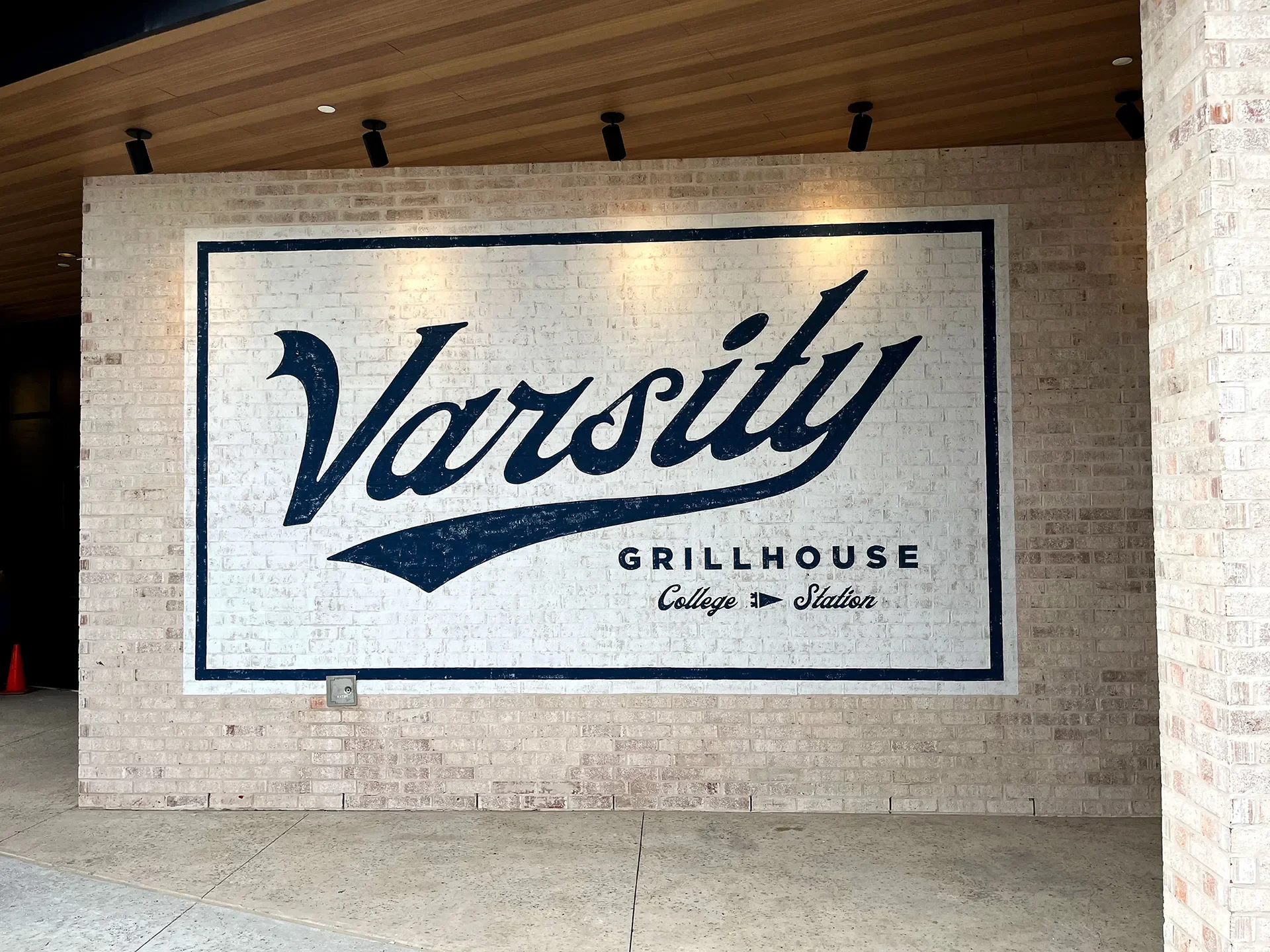 A large sign on a brick wall that reads 'Varsity Grillhouse' with the words 'College Station' underneath, inside a building with a wooden ceiling and track lighting.