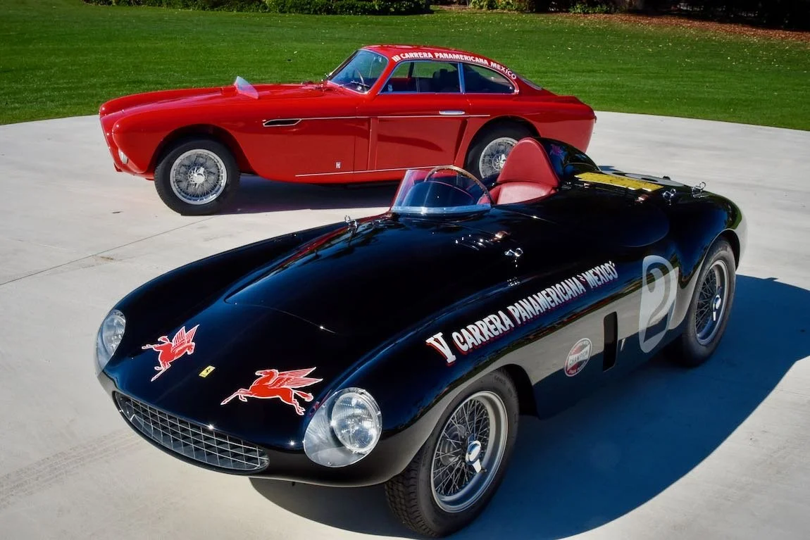 Two vintage racing cars, one black with red and white decals and the other red, parked on a concrete surface with grass in the background.
