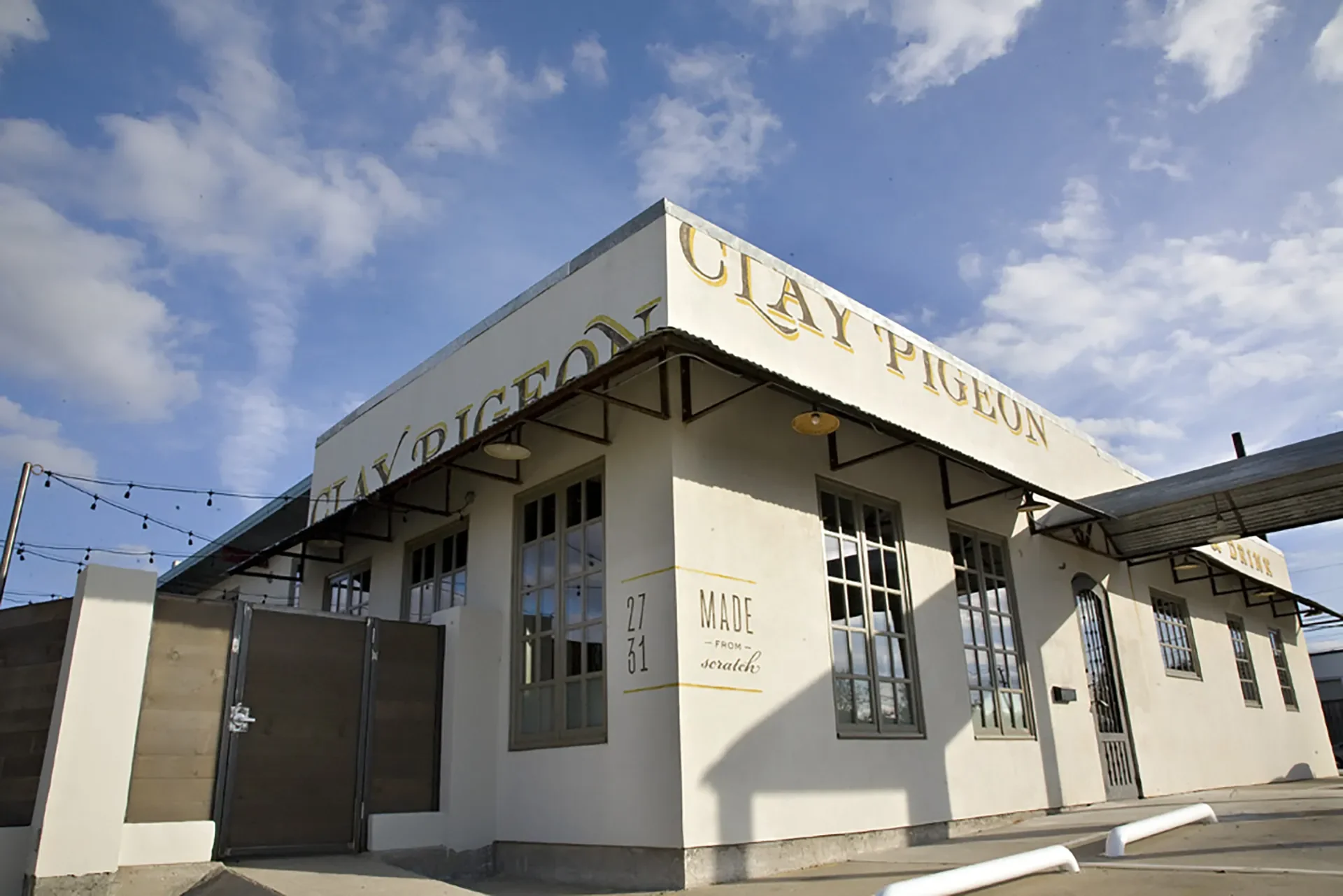 A white building with signs reading 'City of Pigeon' and 'Made from scratch' on the exterior, with multiple large windows and a black gate. Overhead string lights are visible against a partly cloudy blue sky.