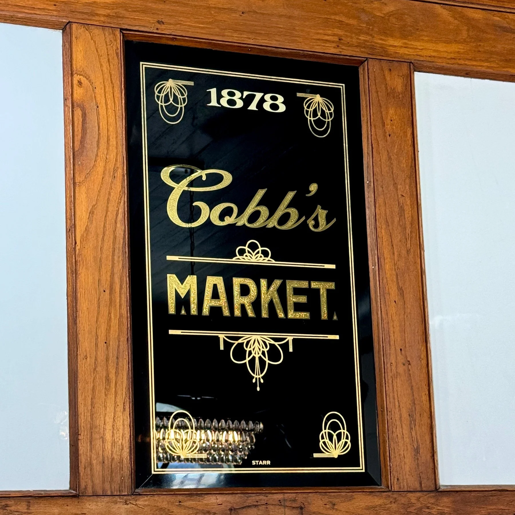 A vintage-style black and gold sign reading 'Cobb's Market,' established in 1878, framed by wood, with reflections of a chandelier visible on the glass.
