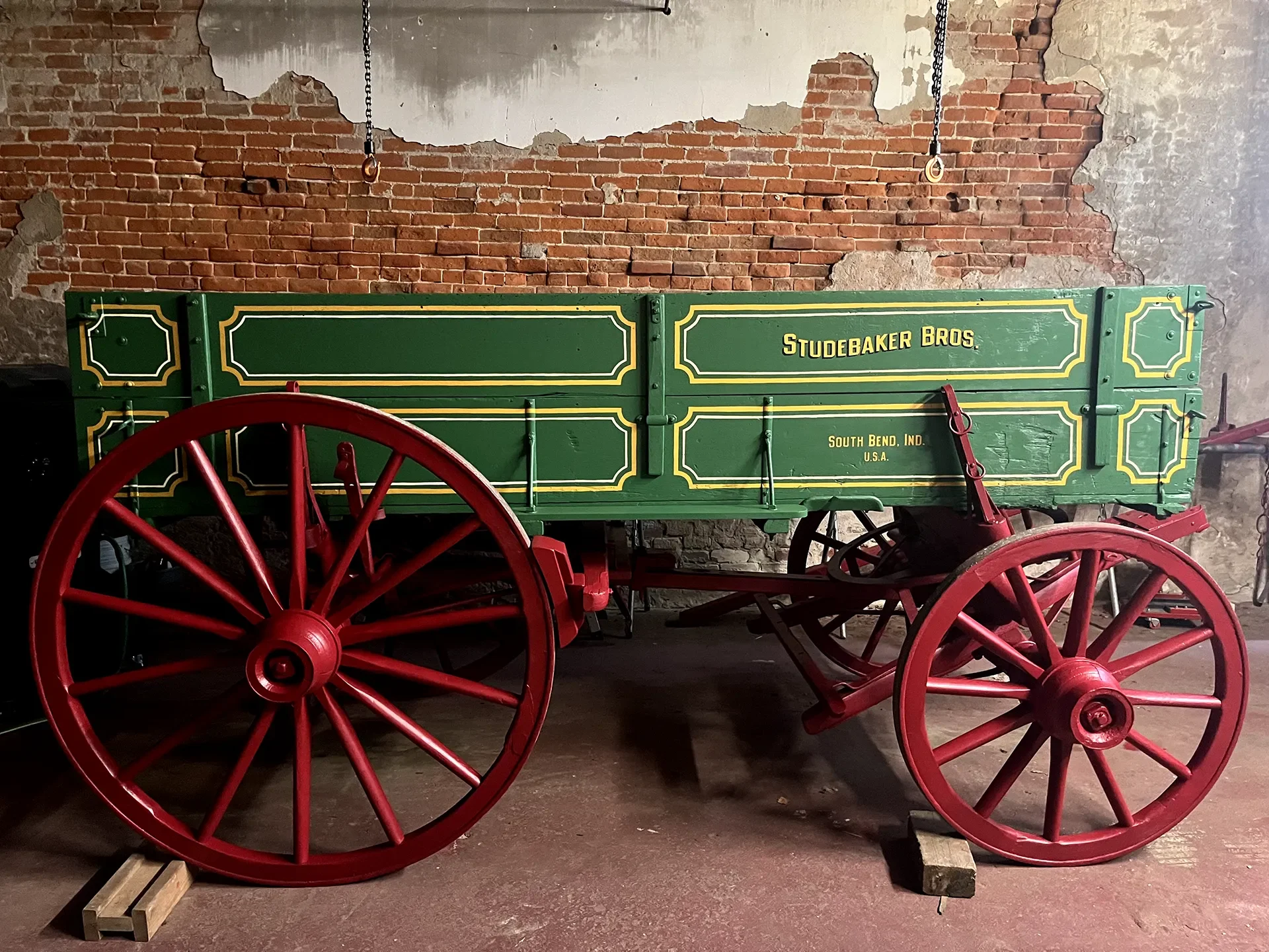 A vintage green wooden wagon with yellow trim, labeled 'Studebaker Bros.' and 'South Bend, Ind., U.S.A.' It has large red wheels and is set against a brick wall with peeling paint.