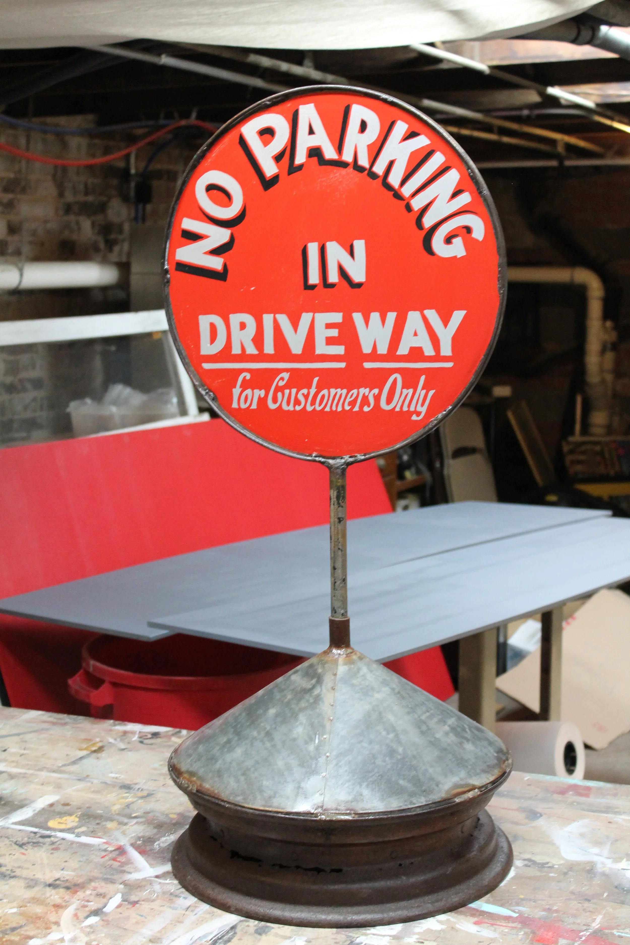 A vintage metal sign with red background and white and black text that says "No Parking in Driveway for Customers Only". The sign is mounted on a rusty stand and is placed on a cluttered work surface in a garage or workshop.