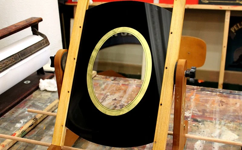 A black circular mirror or frame with a brass oval border, mounted on a wooden stand, in a workshop setting.