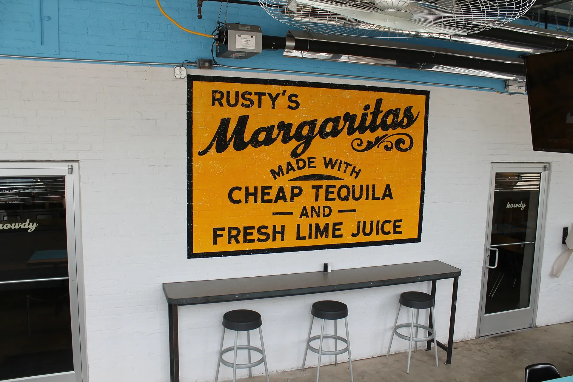 A yellow sign on a white brick wall advertising Rusty's Margaritas made with cheap tequila and fresh lime juice, with a table and three black stools below it.