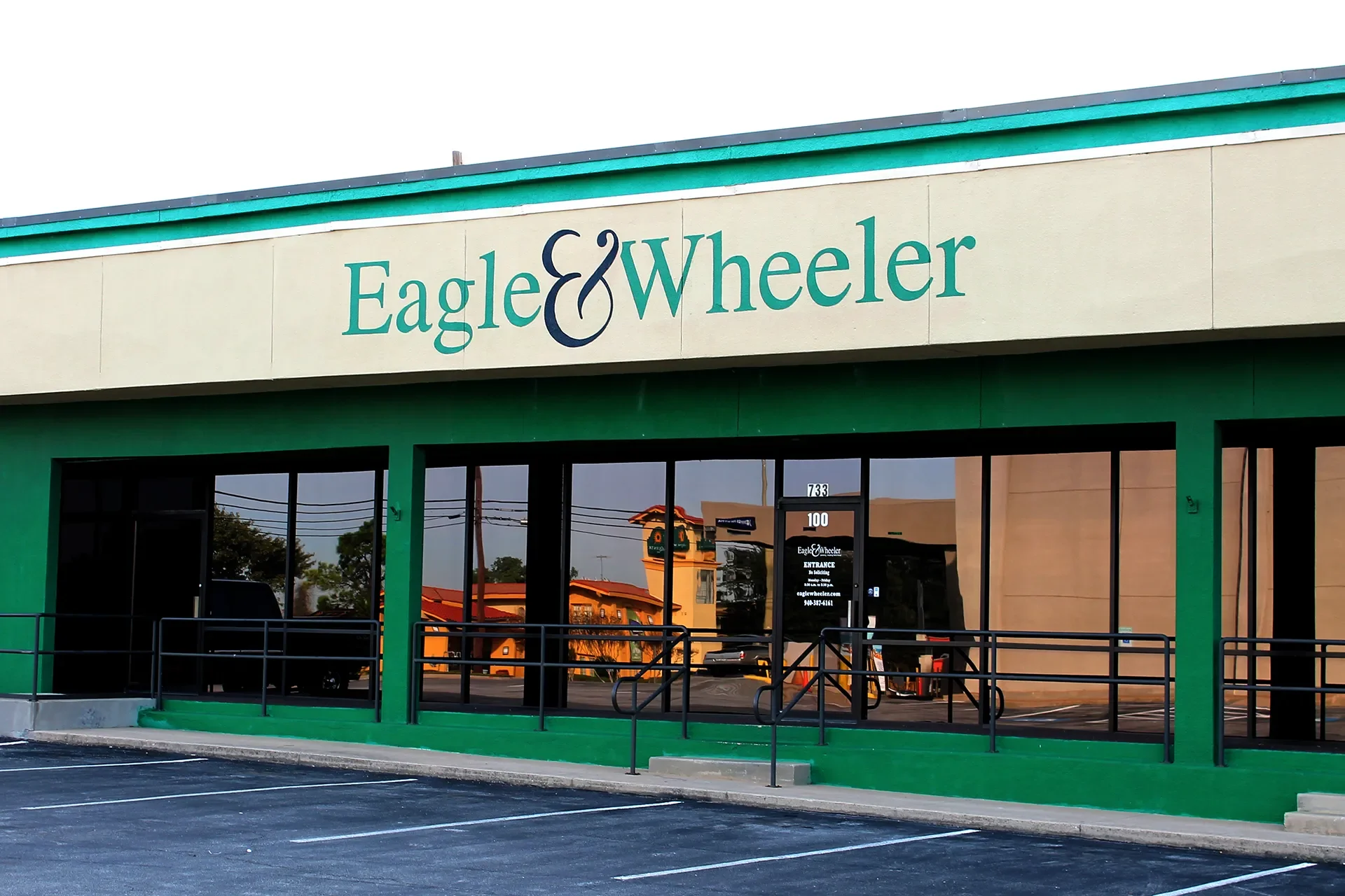 Exterior of a retail store named Eagle & Wheeler, with a beige facade and green accents, large glass windows, a wheelchair accessible ramp, and parking lot in front.