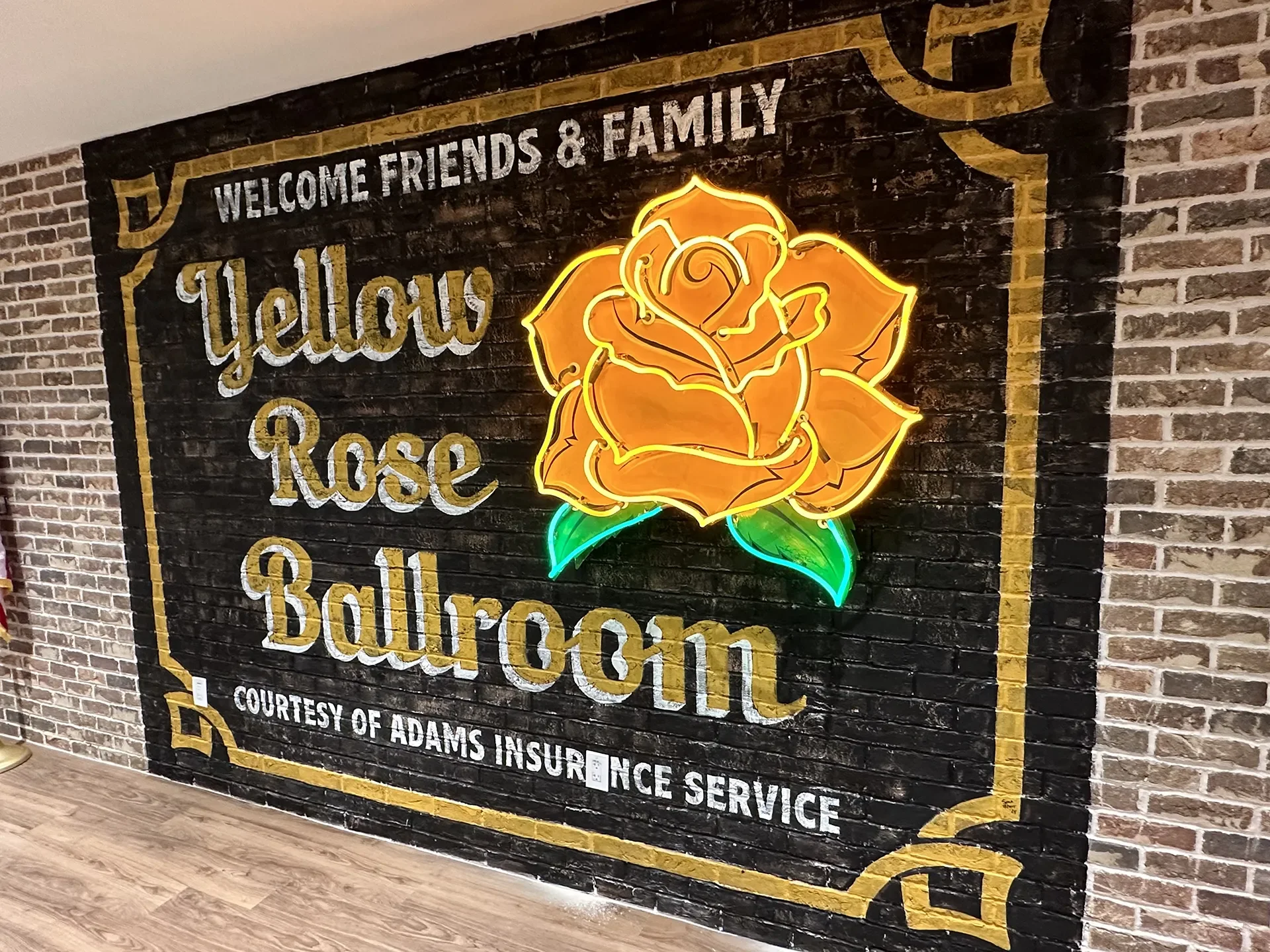 Neon sign with a yellow rose and the text 'Yellows Rose Ballroom' on a black brick wall background, along with a smaller text beneath that reads 'Courtesy of Adams Insurance Service'.
