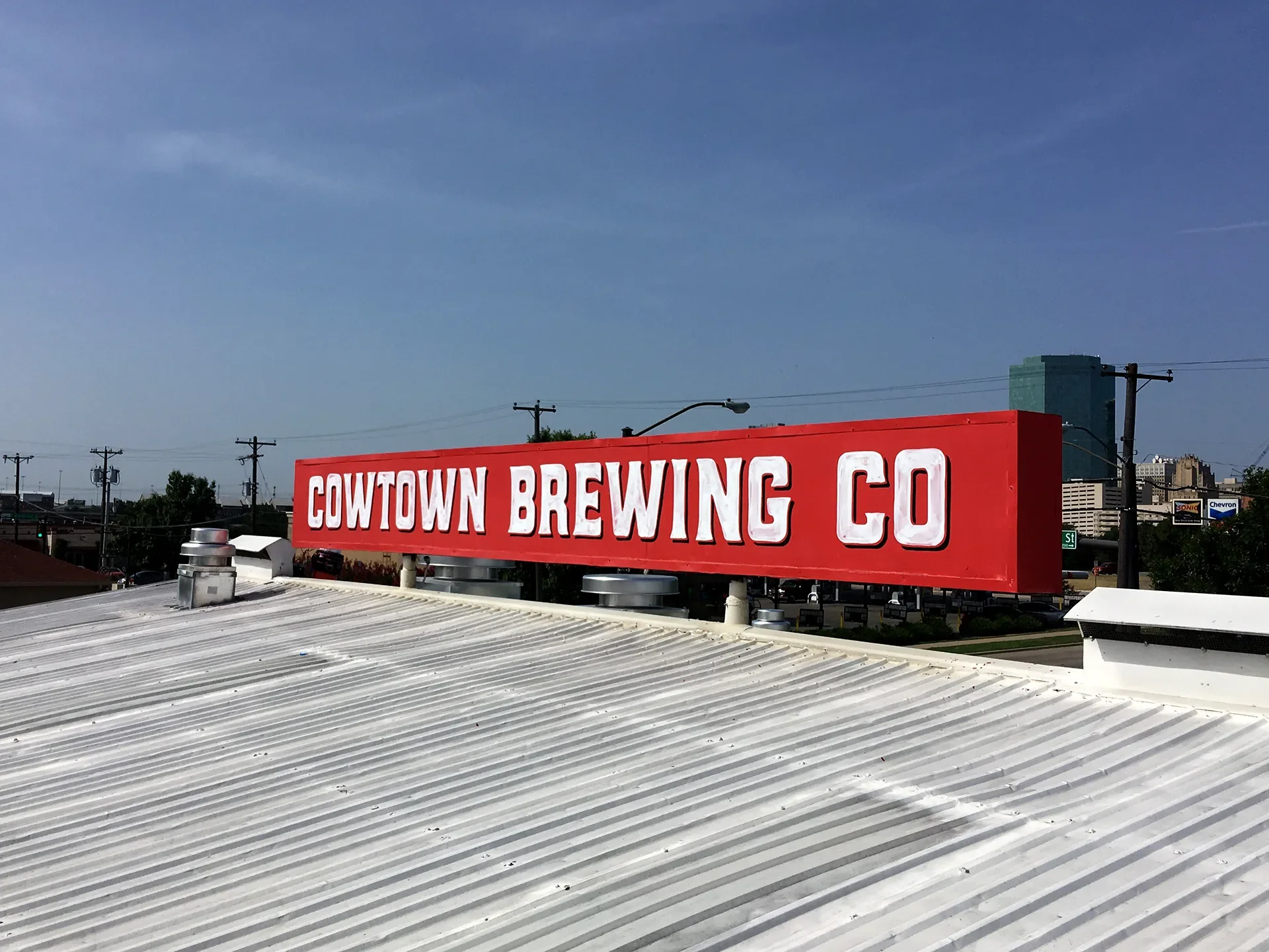 Red signboard on a rooftop with white text that reads 'COWTOWN BREWING CO,' against a partly cloudy sky with a city skyline and power lines in the background.