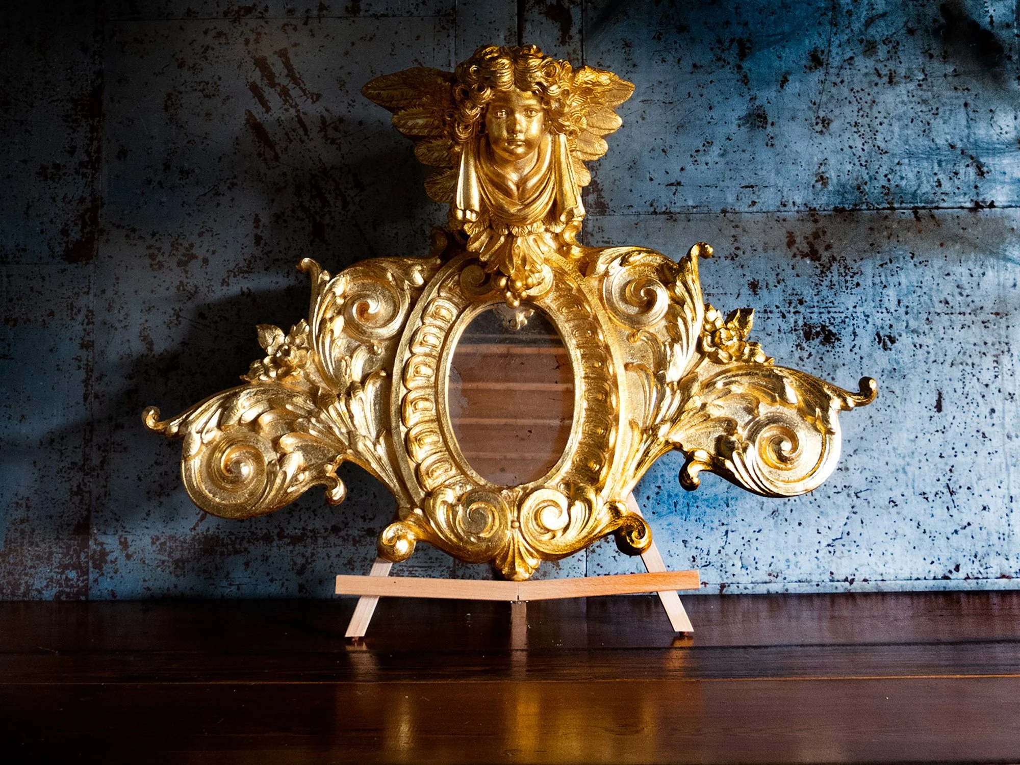 A decorative golden mirror frame with an ornate design, including a human face with wings at the top, set on a small easel on a dark wooden surface, with a distressed blue wall in the background.