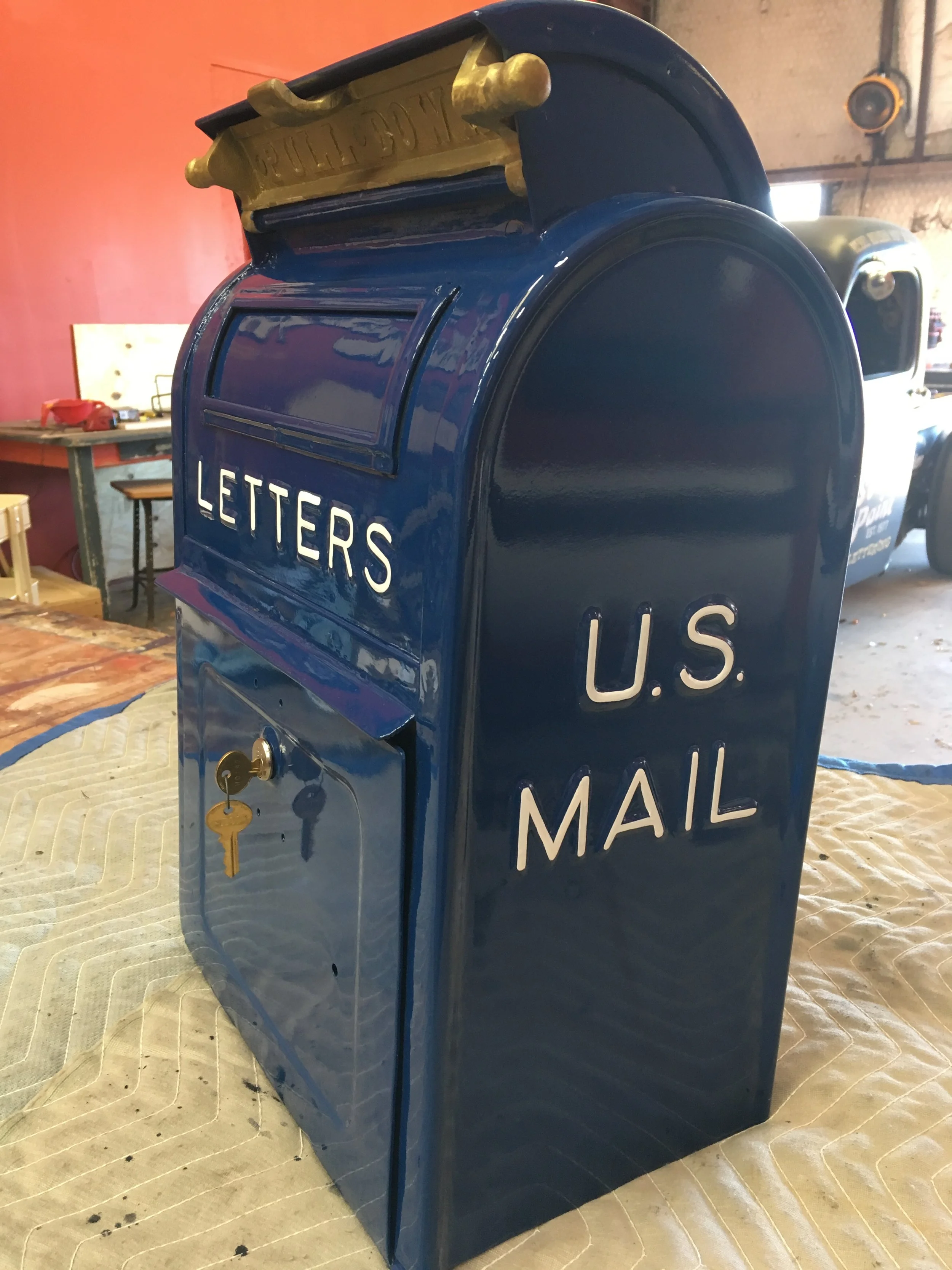 A vintage U.S. Mail letter collection box painted in dark blue with white lettering. The words "LETTERS" and "U.S. MAIL" are visible on the front and side. The box has a slot for letters on the top with a gold-colored decorative trim. A key is insert
