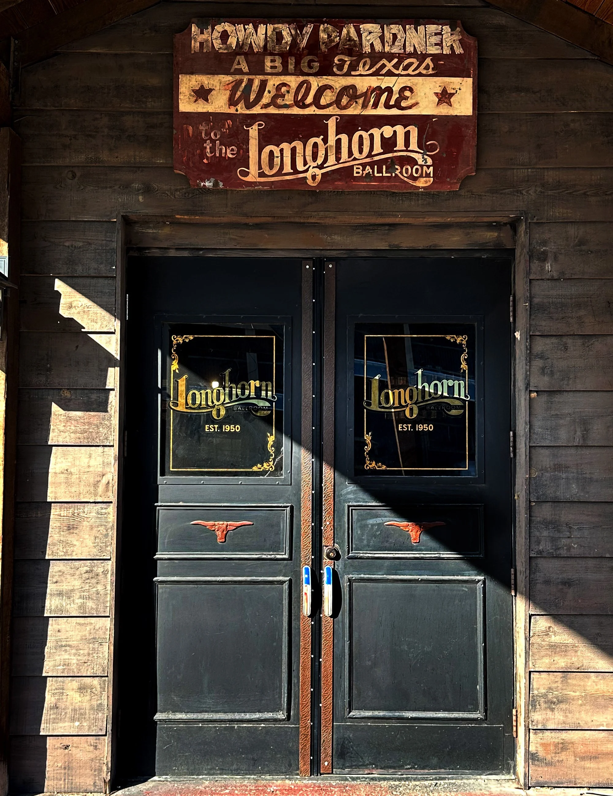 Wooden doors with Longhorn logos and signs indicating 'Est. 1950', leading to the Longhorn Ballroom. Above the doors, a decorative wooden sign welcomes visitors to the Longhorn Ballroom and references a big Texas-style country music scene.