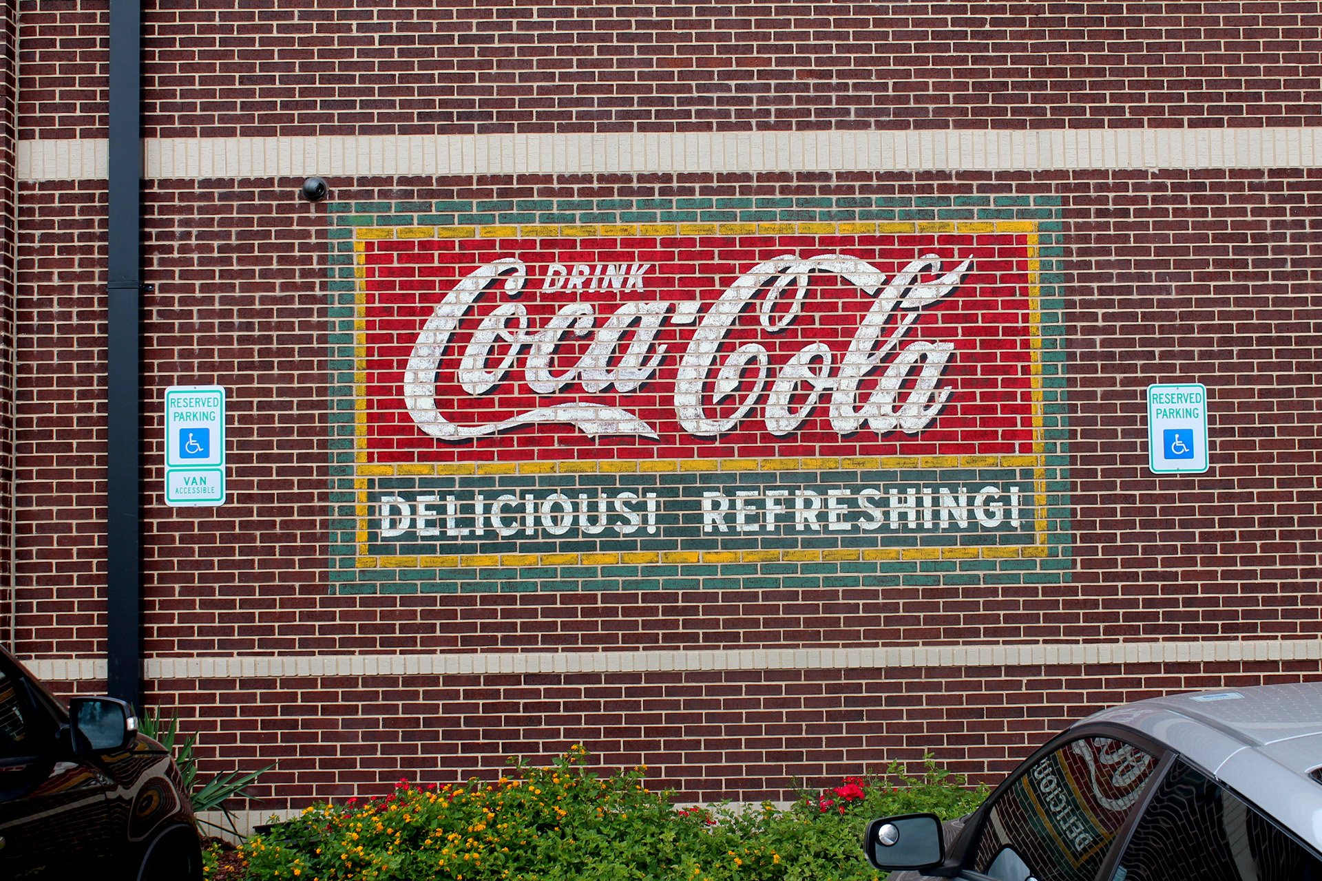 Red brick wall with illuminated Coca-Cola sign reading "Drink Coca-Cola Delicious Refreshing," and two reserved parking signs for handicapped accessible spaces, one on each side of the sign.
