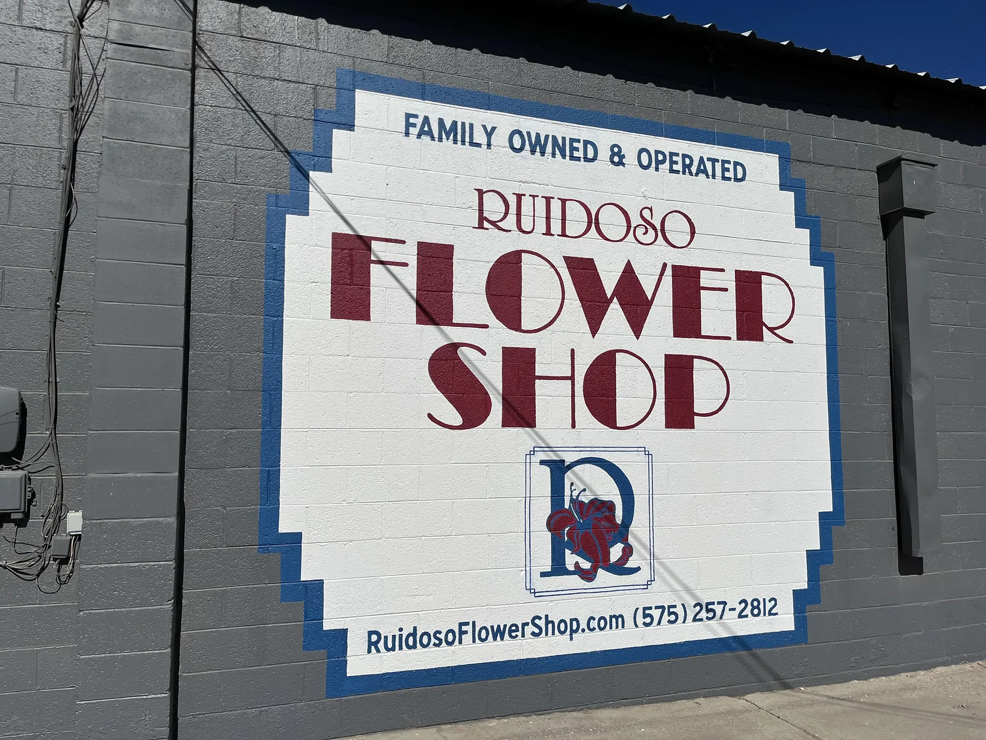 Sign on a brick wall for Ruidoso Flower Shop. The sign states 'Family Owned & Operated Ruidoso Flower Shop' with a logo of a blue letter 'R' and a red flower, and the website RuidosoFlowerShop.com along with the phone number (575) 257-2812.