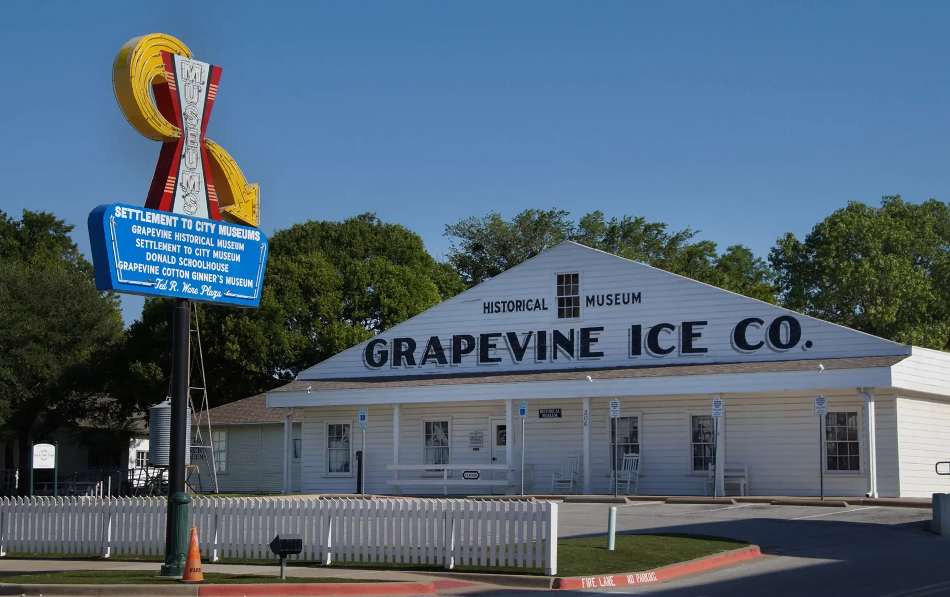 White building with black letters reading "Grapevine Ice Co." and a sign with blue background listing historic museum sites in Grapevine, Texas, with a vintage billboard sign on a pole nearby, greenery and trees in the background, and a white picket 