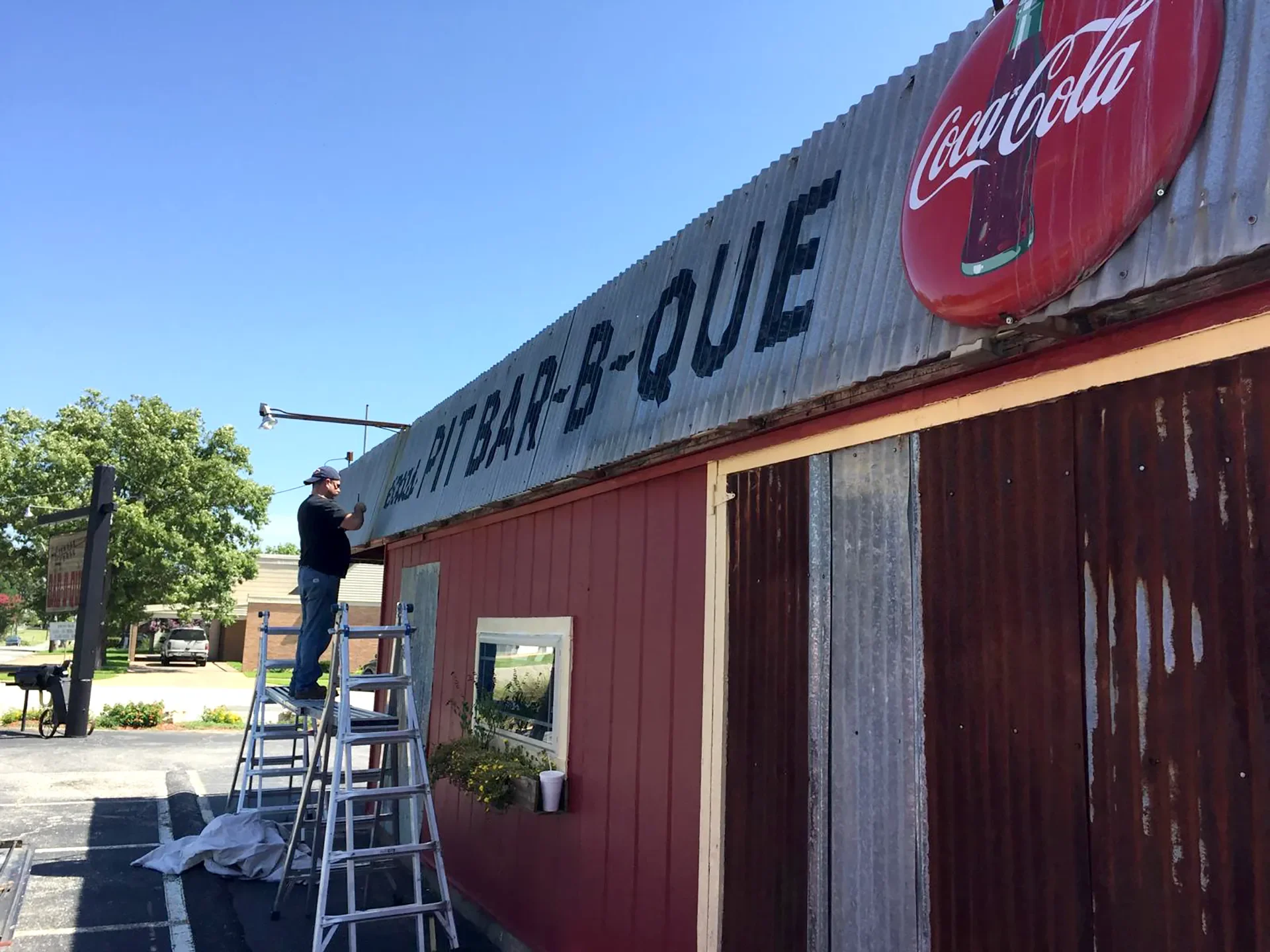 A man standing on a ladder cleaning the signboard of a restaurant called 'Bite-B Que' with a Coca-Cola logo on the side. The building has a red and metal exterior with a small window with flower boxes underneath. There are trees and cars in the backg