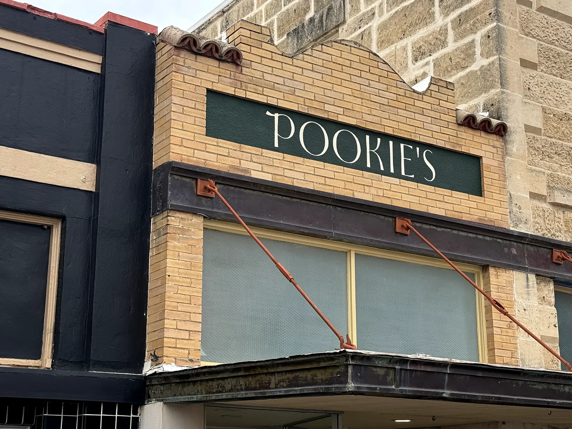 Close-up of a building exterior with a sign that reads 'POOKIE'S' mounted on a brick wall above large window with a black awning and orange support bars.