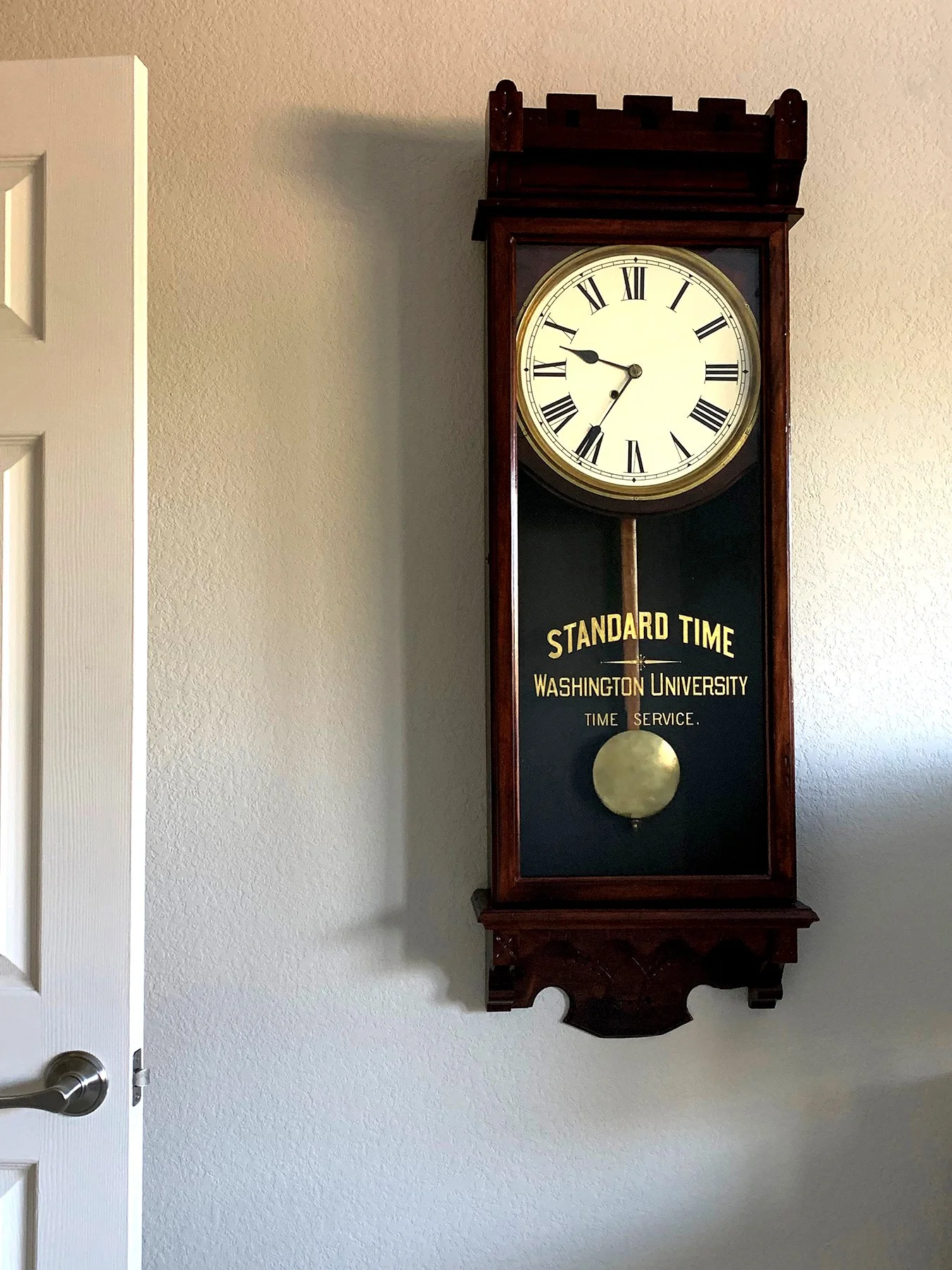 A vintage wall clock with a round face, Roman numerals, and a pendulum, mounted on a beige wall near a white door. The clock has a dark wood frame and displays the time as 10:47.