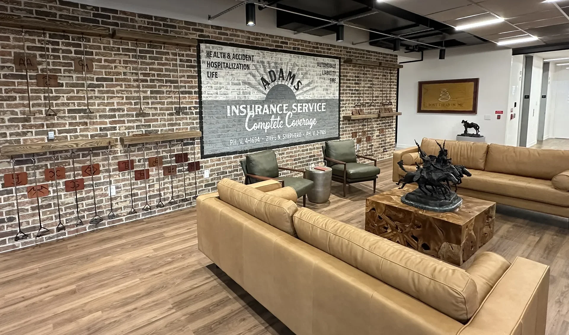 A lobby area with exposed brick wall, furnished with leather sofas, two green armchairs, a wooden table with a horse sculpture, and a large sign for Adams Insurance Service on the brick wall. There is a framed picture on the white wall and a small ho