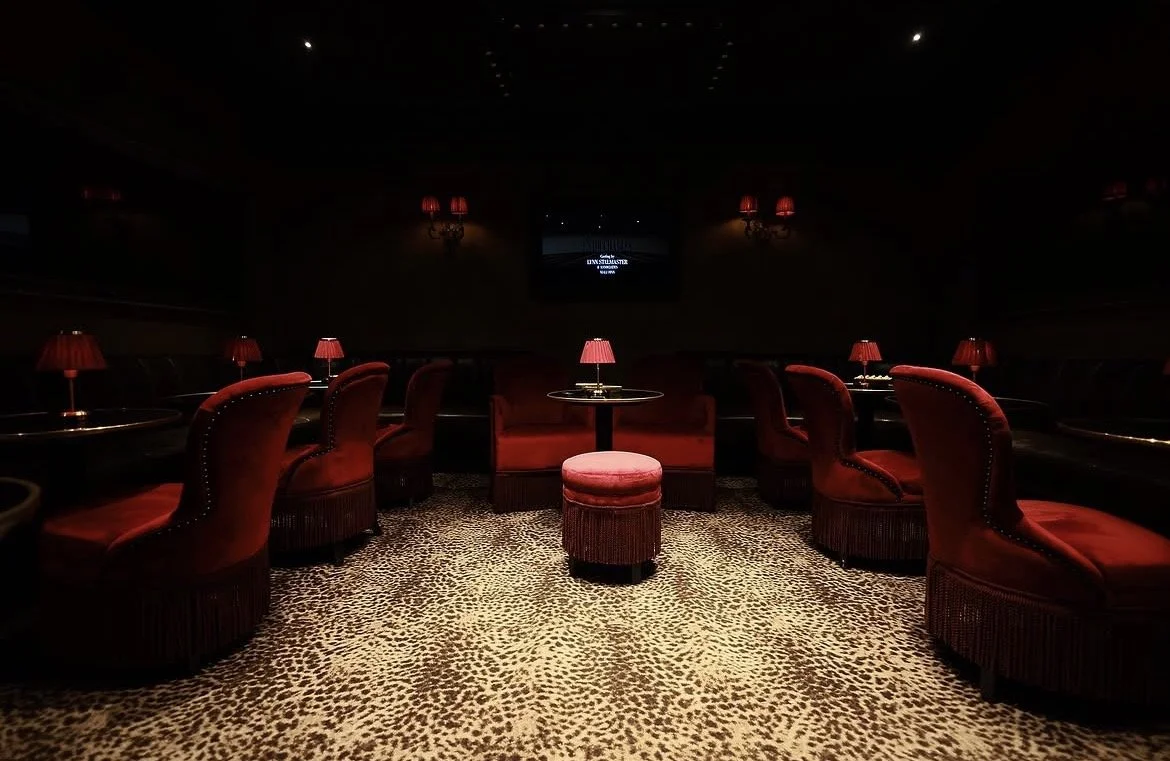 Salle de cinéma privée avec dim lighting, fauteuils en velours rouge, petites lampes de table, tapis à motifs léopard, écran au fond.