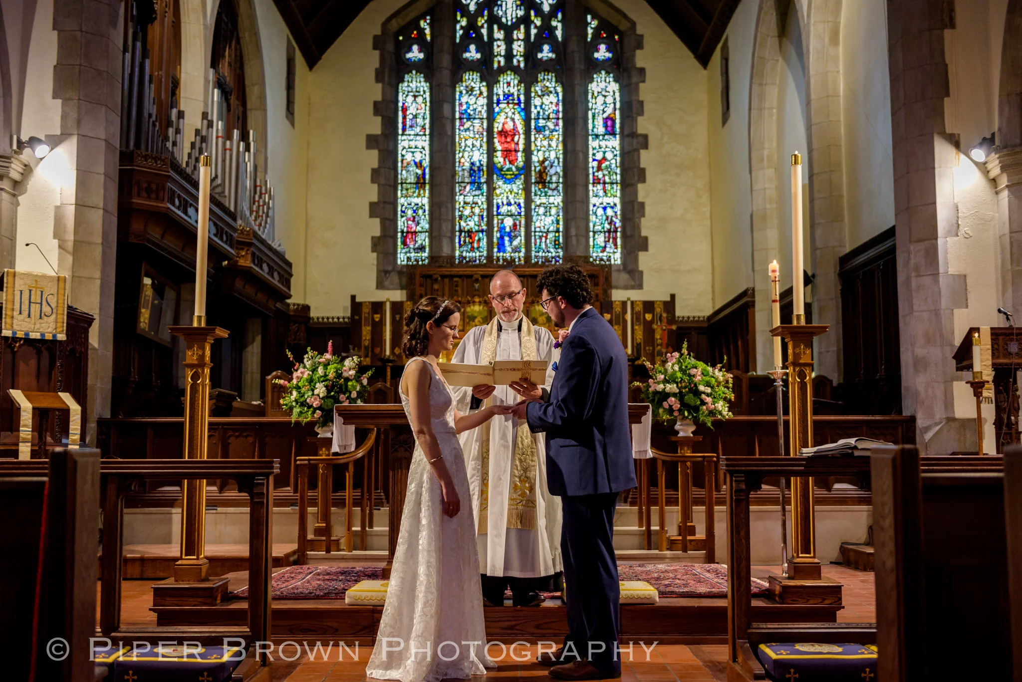 Slim brunette bride in a white embroidered gown exchanging rings with a Curley haired brunette groom in a navy suit in front of a priest in a church with ornate stain-glass window behind them.