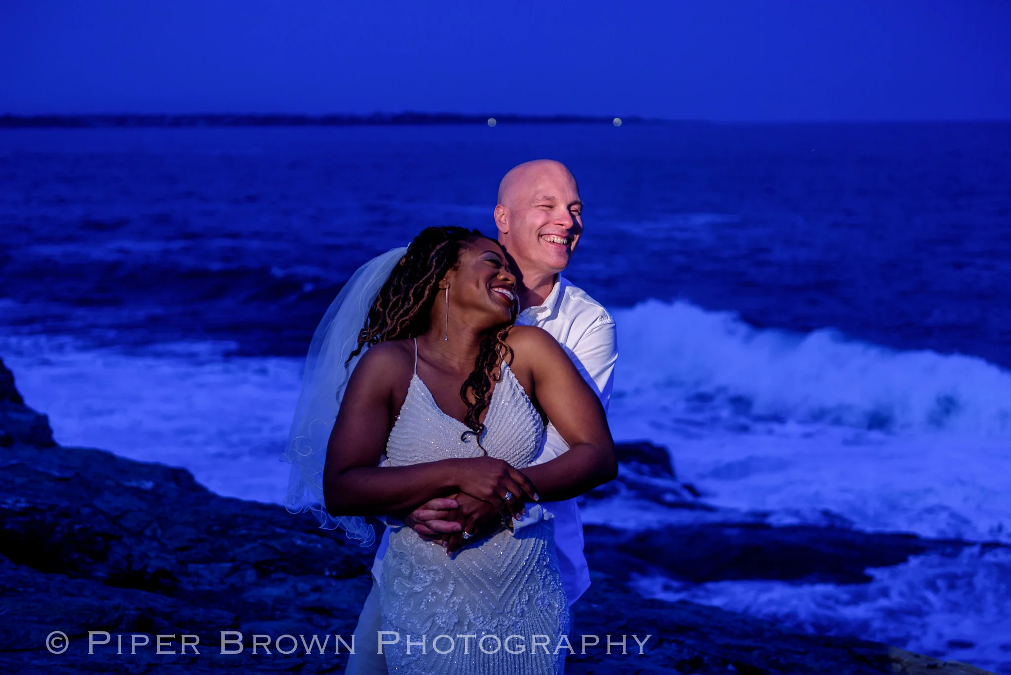 Black smiling woman in a beaded shoe-string wedding dress embraced by laughing white bald man with mean surf behind them.