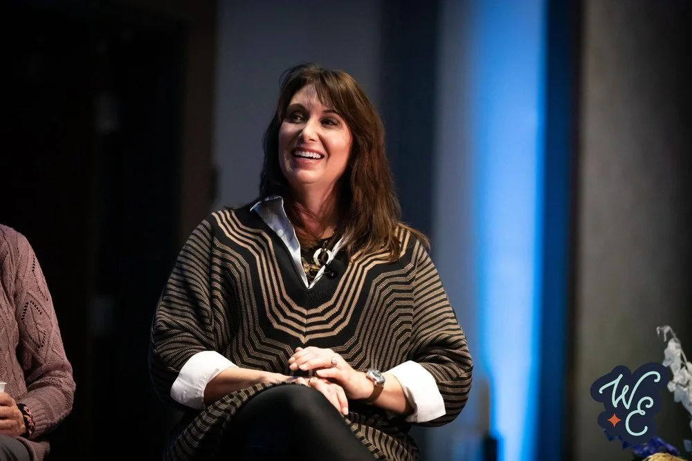 A woman with long brown hair, smiling, wearing a patterned sweater over a white shirt, sitting on a chair with a dark background and a blue light to her right.