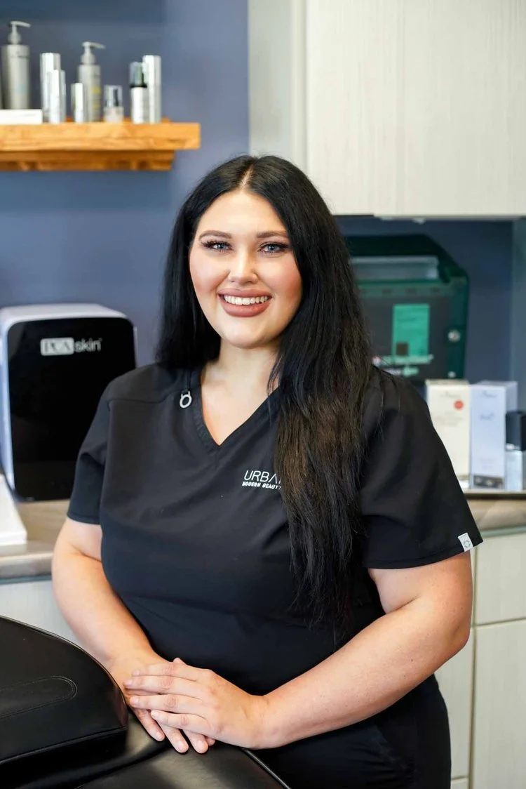 A smiling woman with long dark hair and light skin in a black uniform standing in a spa or clinic setting.