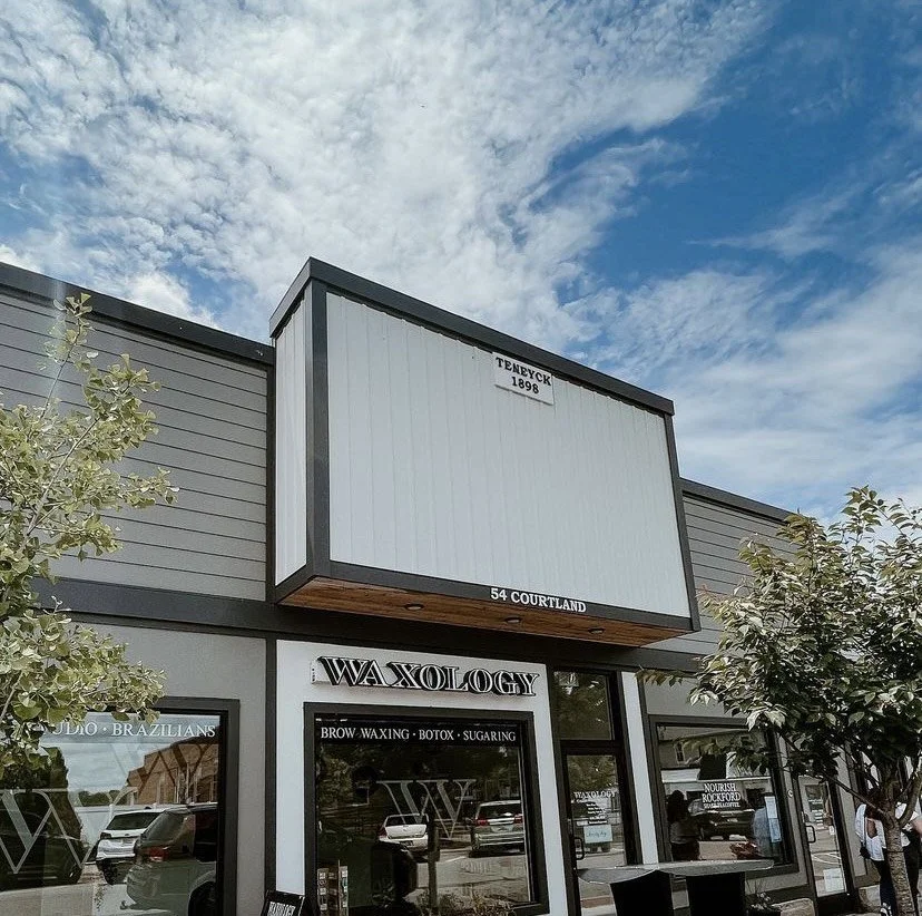 Exterior view of a building with a large, empty signboard above the entrance. The building has storefront windows with signs for a salon offering brow waxing, Botox, and sugaring. There are trees and parked cars outside, and a partly cloudy sky overhead.