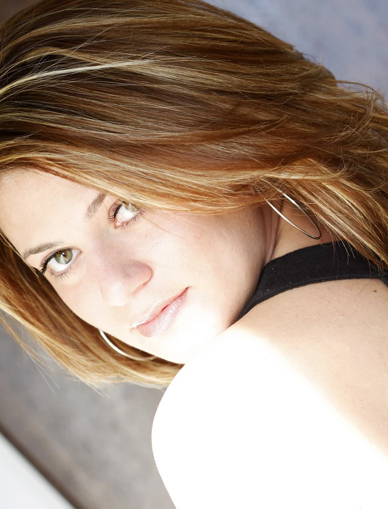 Close-up of a woman with shoulder-length reddish-brown hair, green eyes, and hoop earrings, looking over her shoulder.