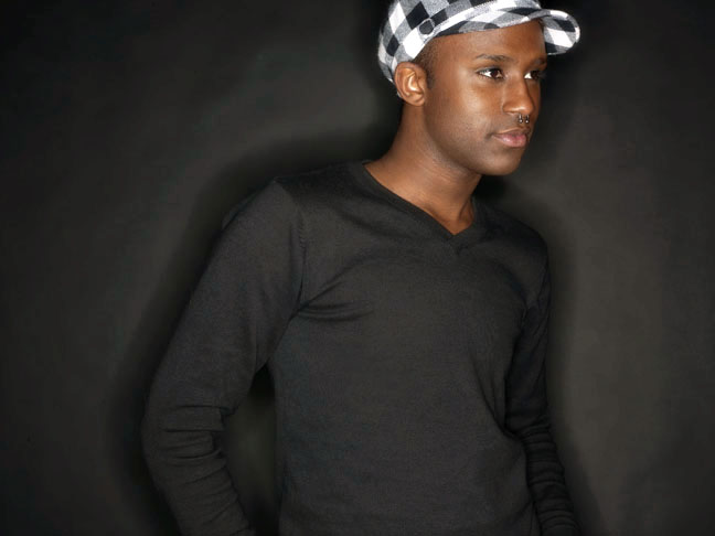 Young man wearing a black long-sleeve shirt and a black and white checkered cap, standing against a dark background.