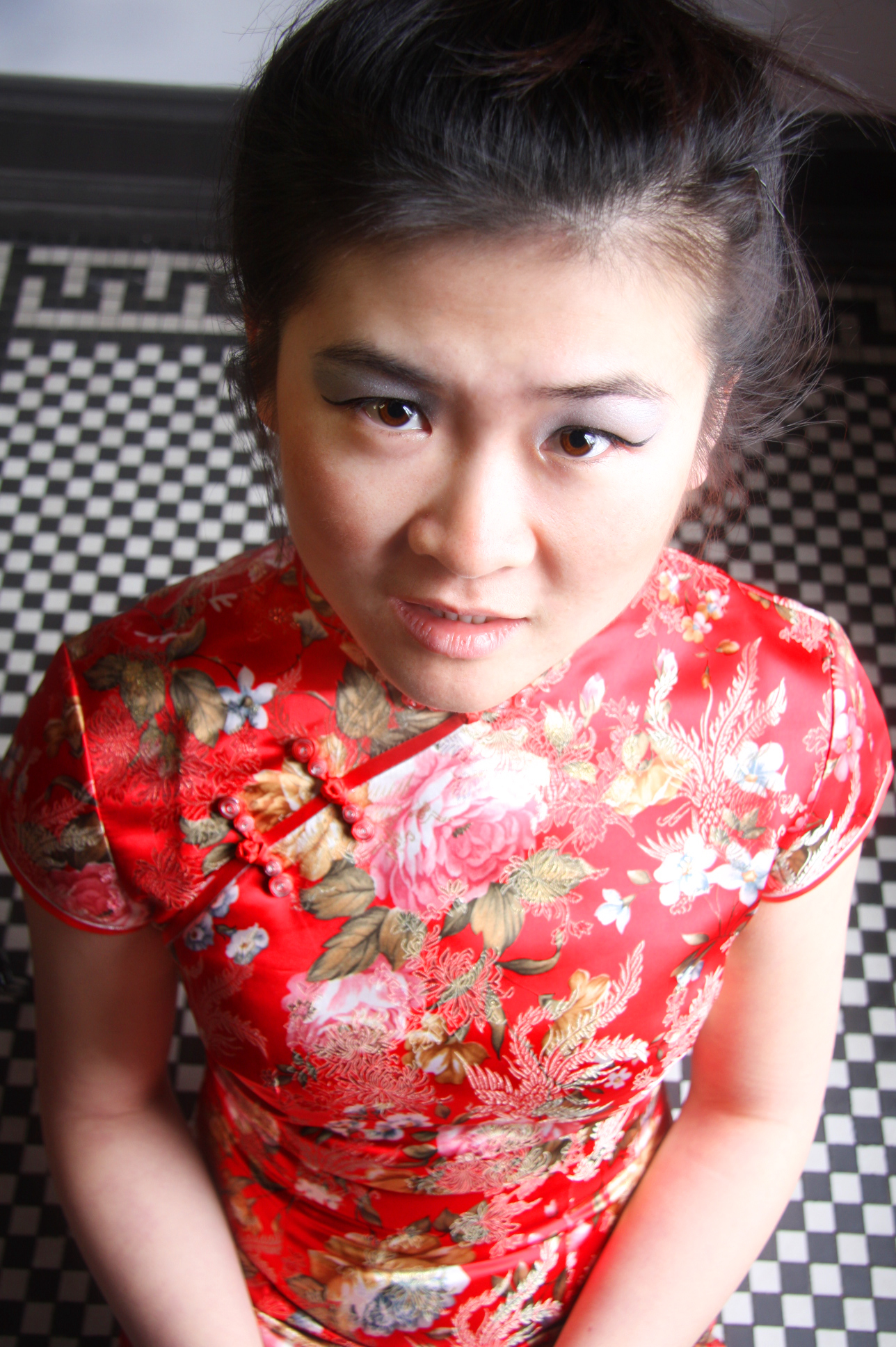A woman wearing a red floral cheongsam dress, looking up at the camera with a slight smile, standing on a black and white checkered floor.