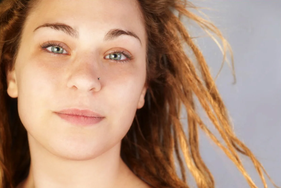 Close-up of a woman with red dreadlocks, light skin, and a nose piercing, gazing at the camera with a slight smile.