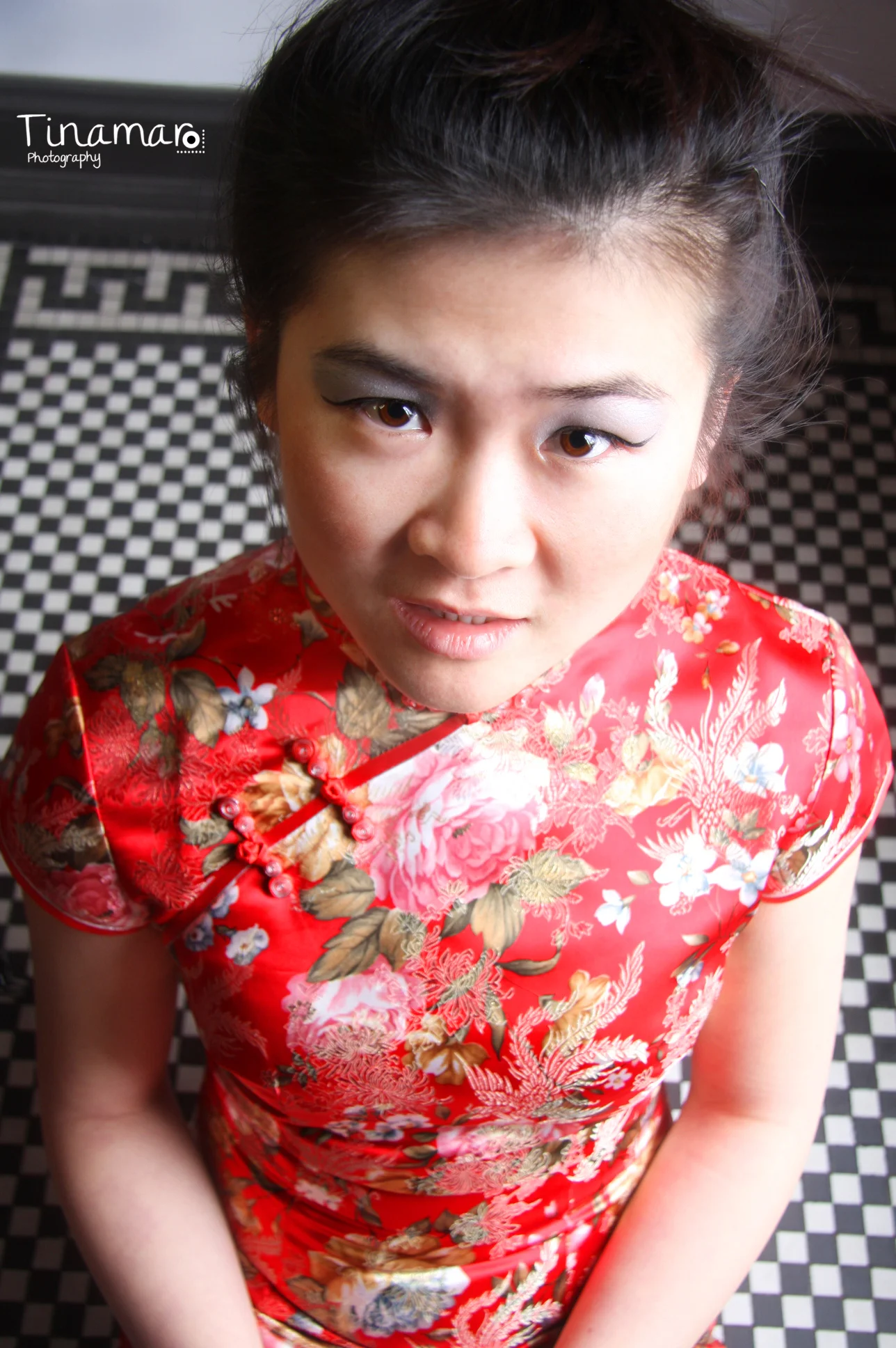 A woman looking up at the camera while wearing a red floral traditional dress, standing on a black and white checkered floor.