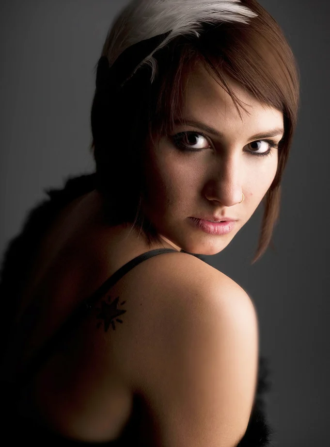 Close-up portrait of a young woman with short brown hair and a white feather in her hair, looking over her shoulder at the camera against a dark background.