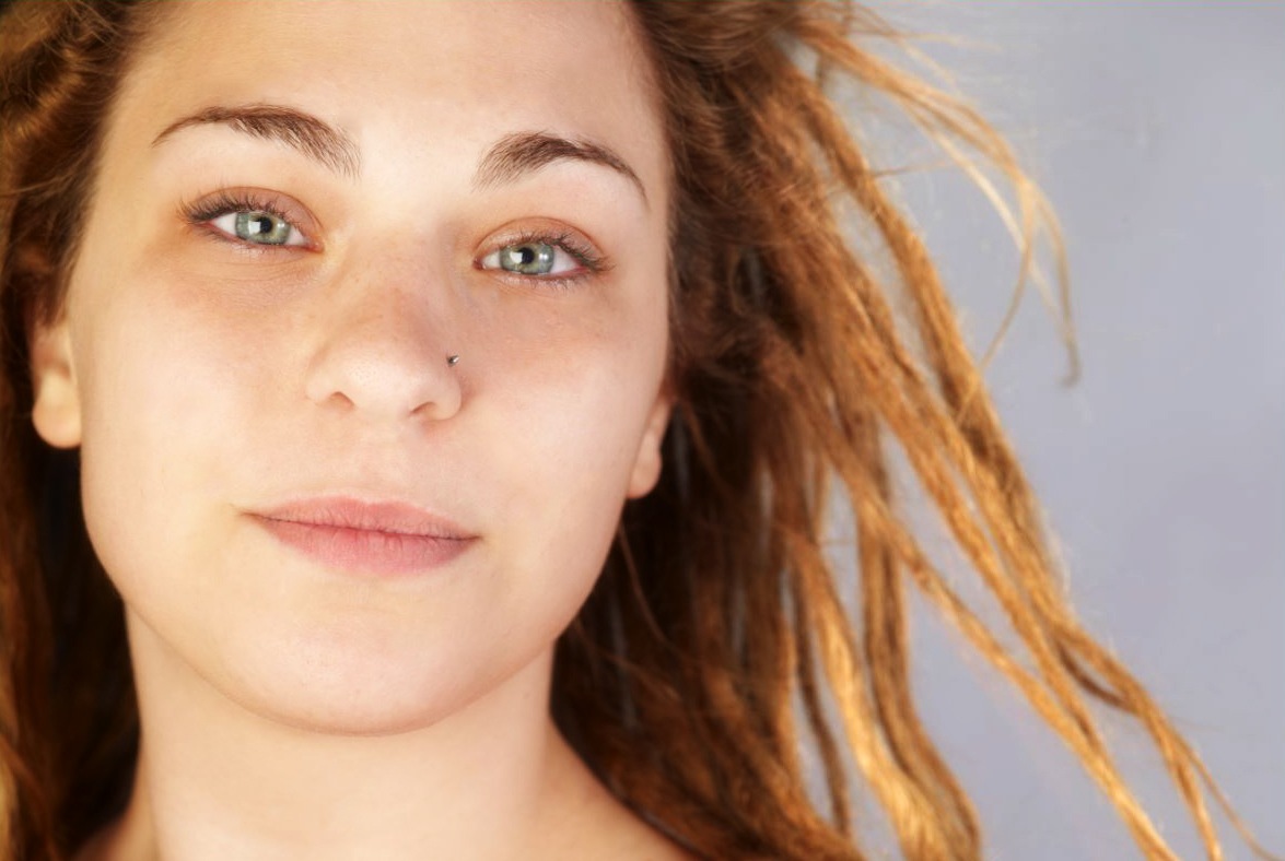 Close-up of a young woman with red dreadlocks and a nose piercing, looking at the camera with a slight smile.