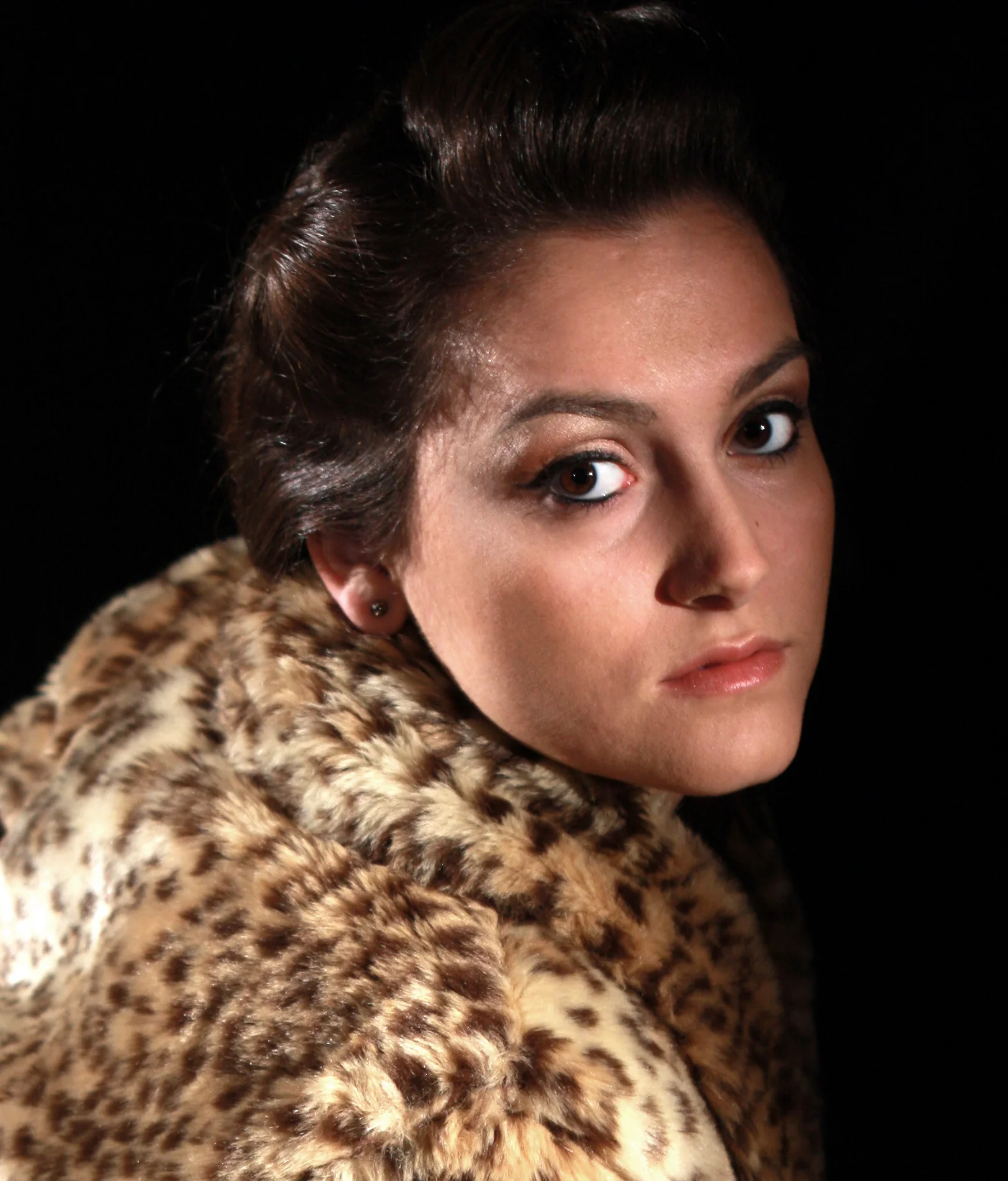 A woman wearing a leopard print coat with short, dark hair styled back, gazing at the camera against a black background.