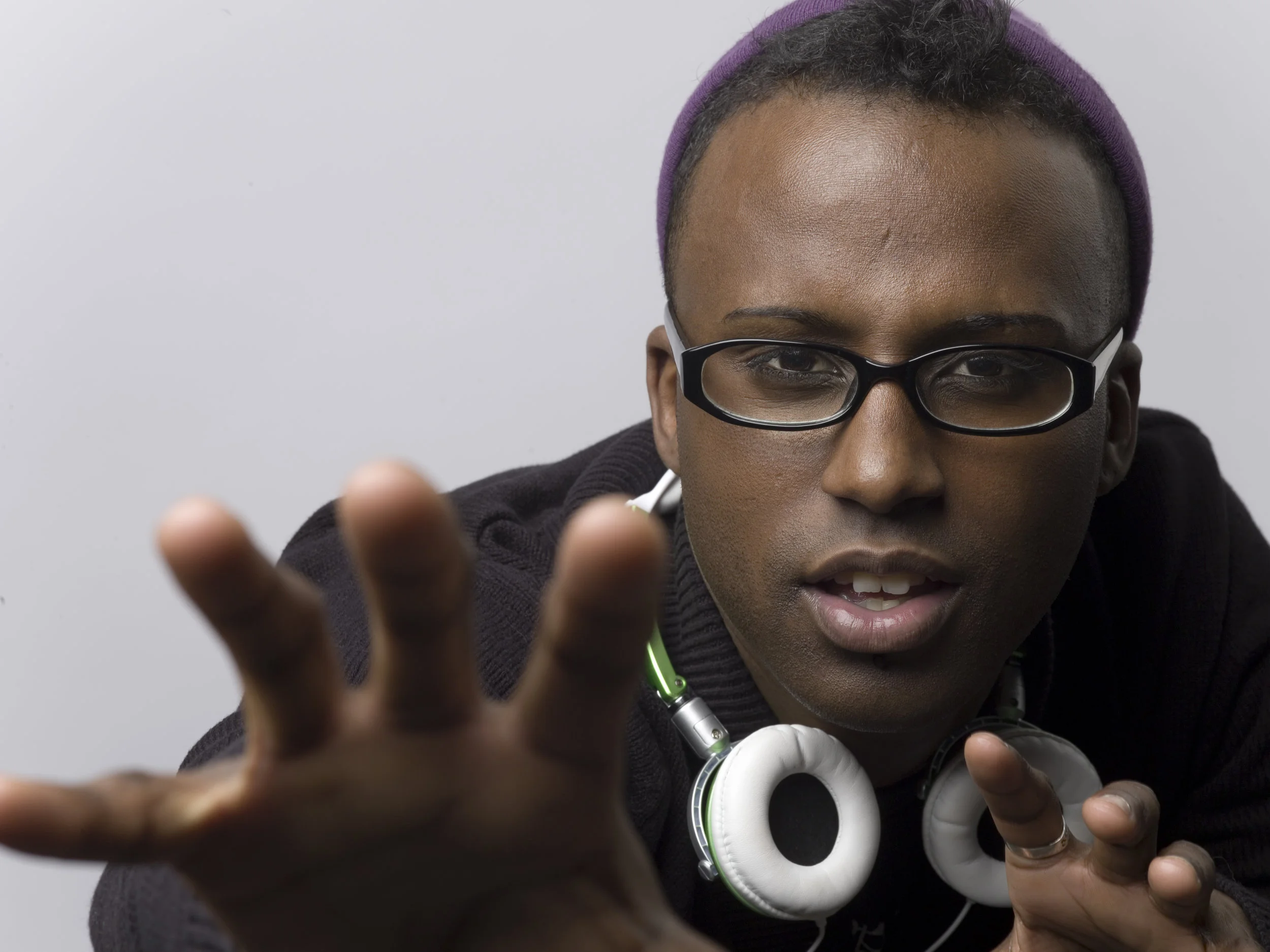 A young man with glasses and a purple beanie reaching towards the camera, with a serious expression, wearing headphones around his neck and a black hoodie.